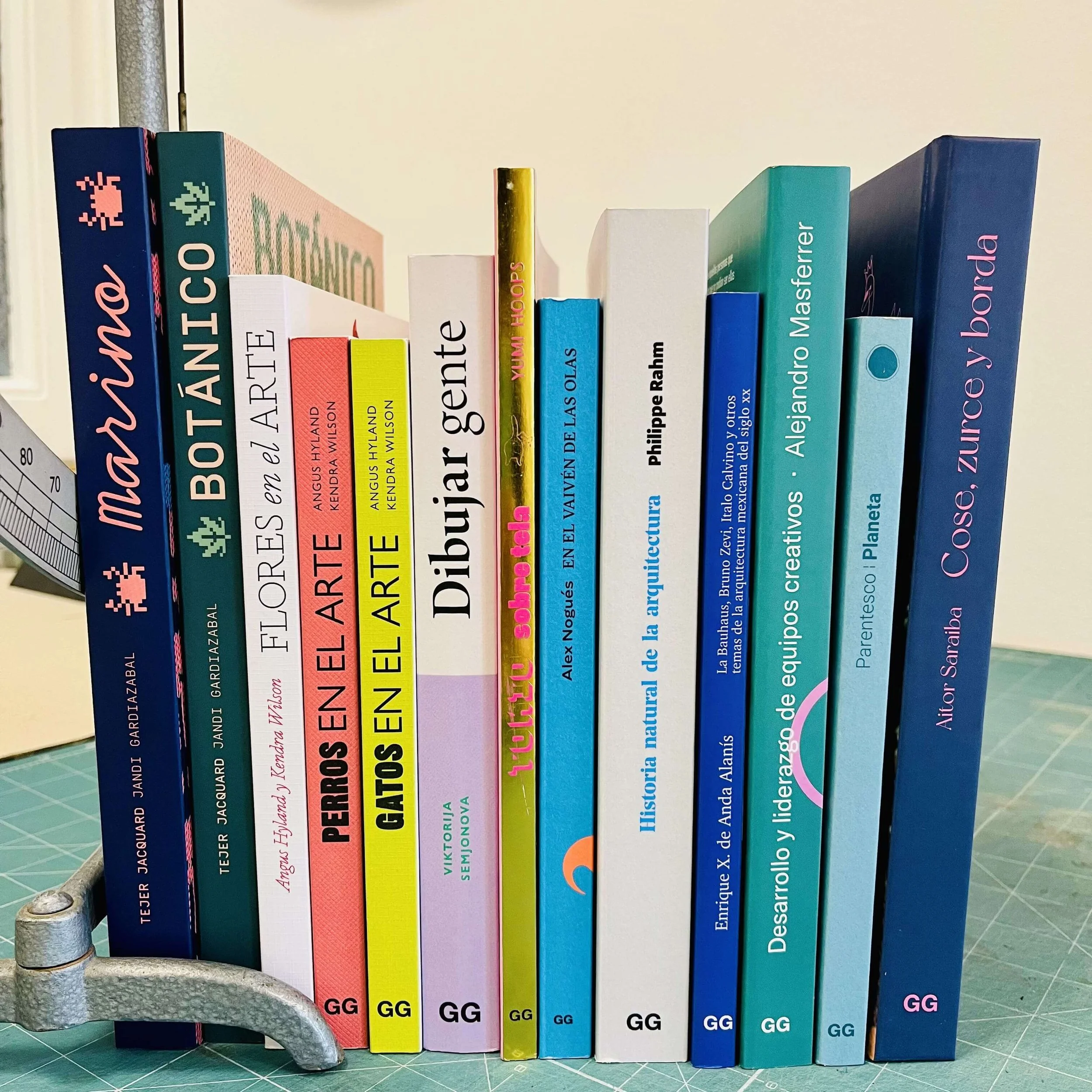 Una pila de libros con varios títulos en español y en inglés, organizados en fila, sobre una mesa con una regla y otras herramientas. Los libros cubren temas de arte, arquitectura, animales, naturaleza, ciencia y desarrollo personal.