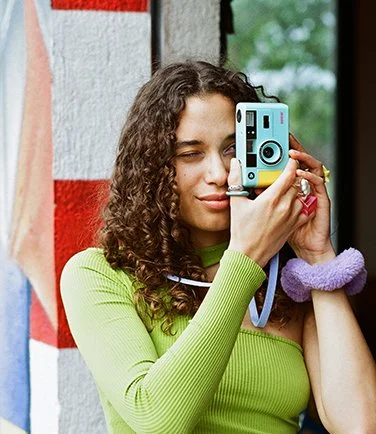 Mujer con cabello rizado sosteniendo una cámara de fotos, usando un suéter verde y una pulsera lila.