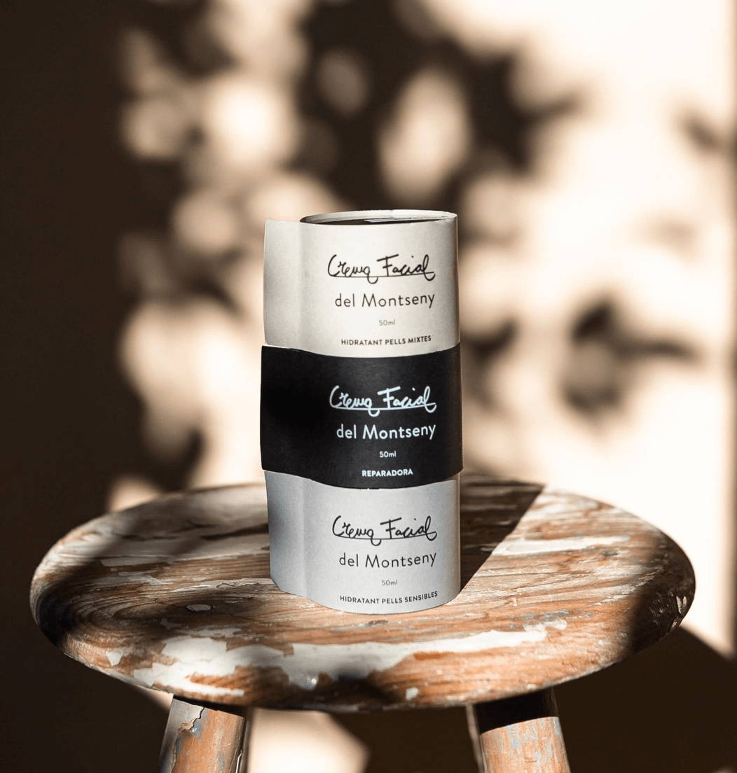 Un envase cilíndrico de crema facial del Montseny, con etiquetas blancas y negras, colocado sobre un taburete de madera envejecido, con un fondo difuso de formas borrosas y luz suave