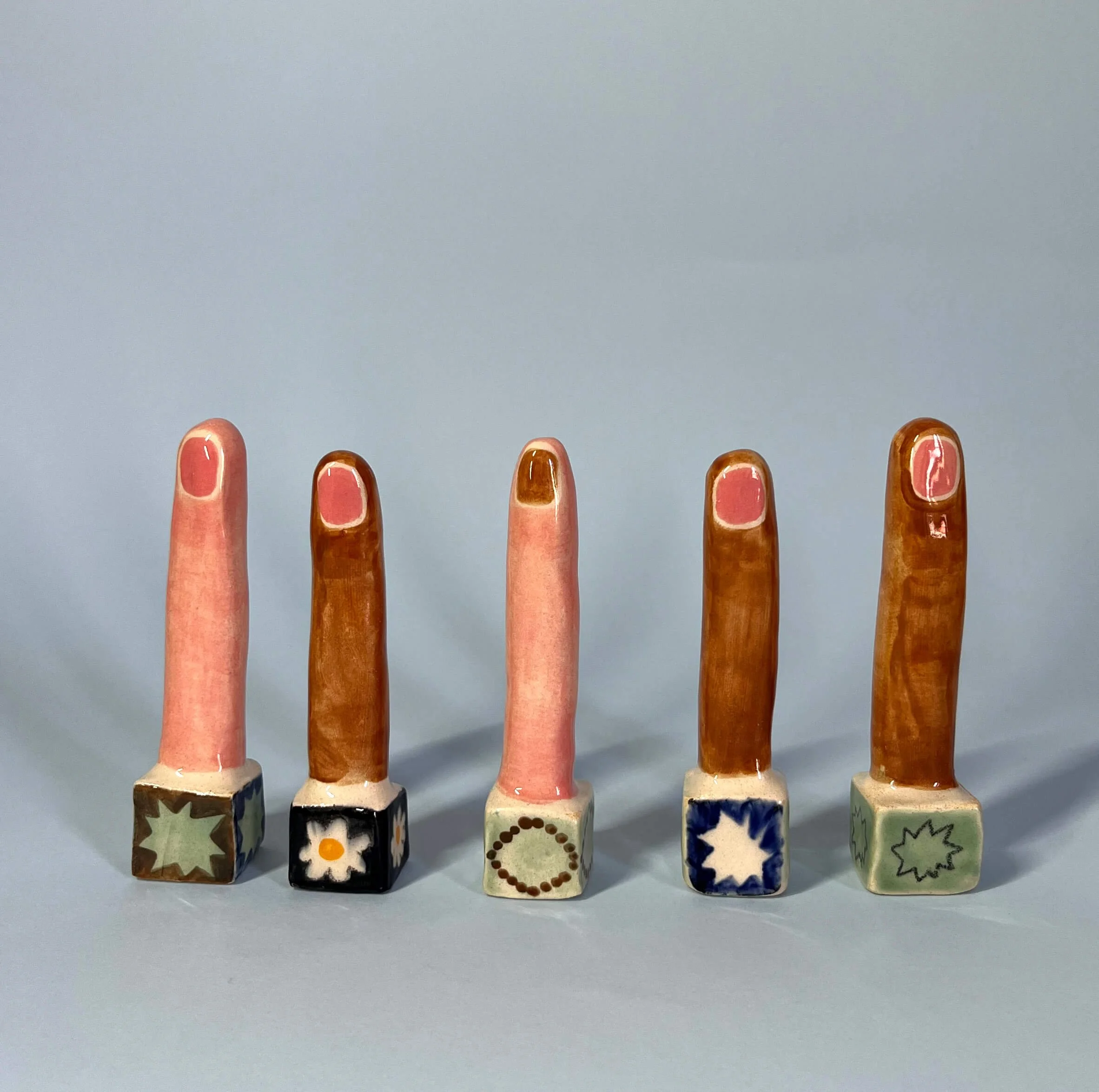 Cinco pequeñas esculturas de dedos con uñas pintadas, sobre bases decoradas, en diferentes tonos de piel y uñas de diferentes formas y colores.