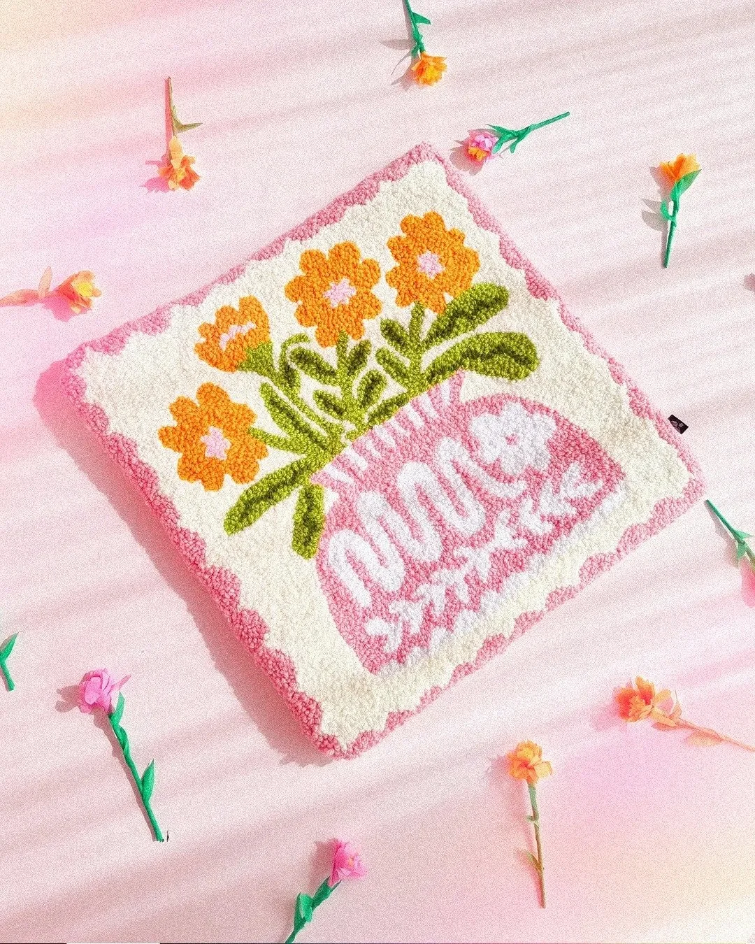 Una toalla decorativa con flor de alhelí y la palabra "DULCE SABOR" bordada, rodeada de pequeñas flores de papel en un fondo rosa.