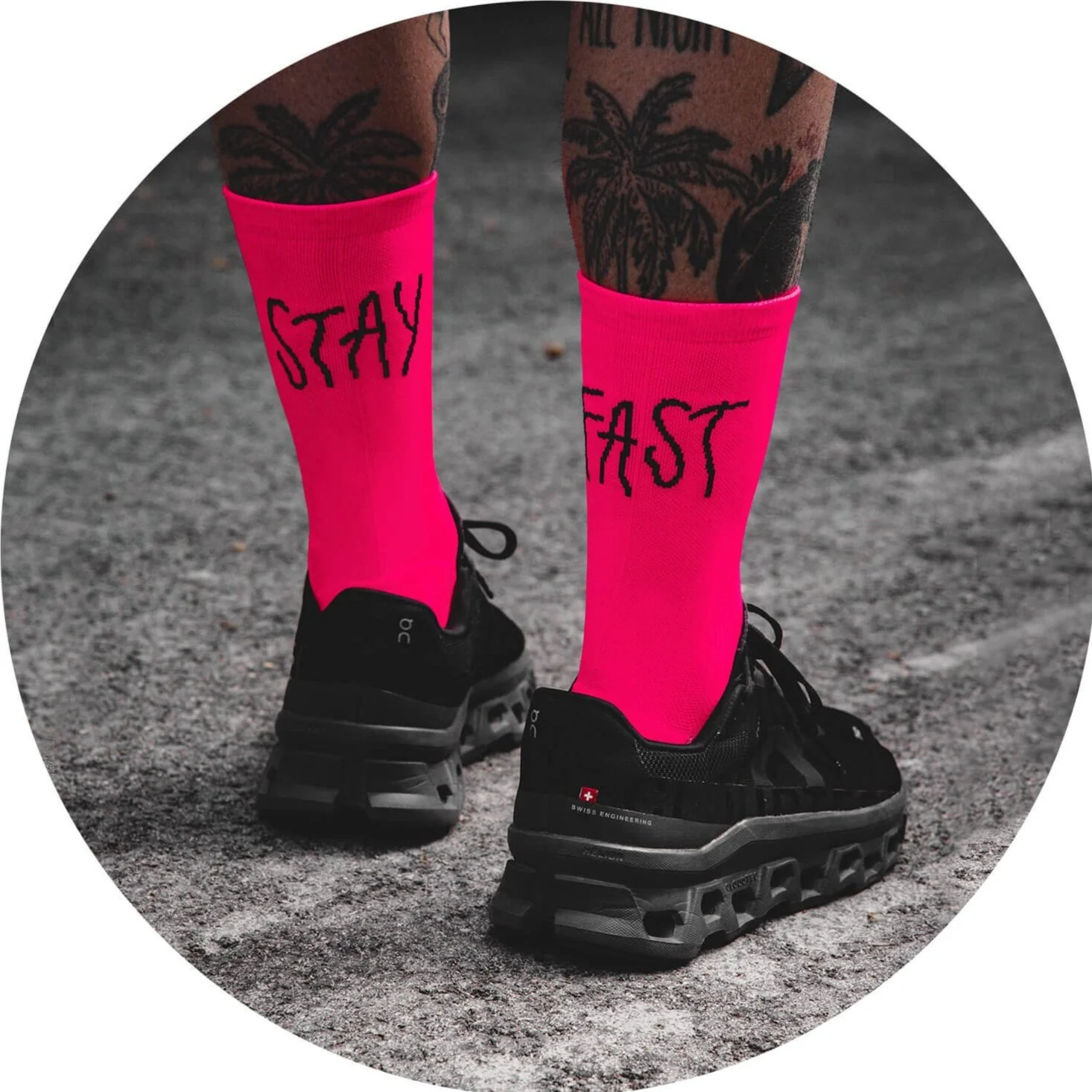 Persona con tatuajes en las piernas usando medias rosas con la palabra "STAY" en una pierna y "FAST" en la otra, y zapatillas negras en una superficie de piso áspero.
