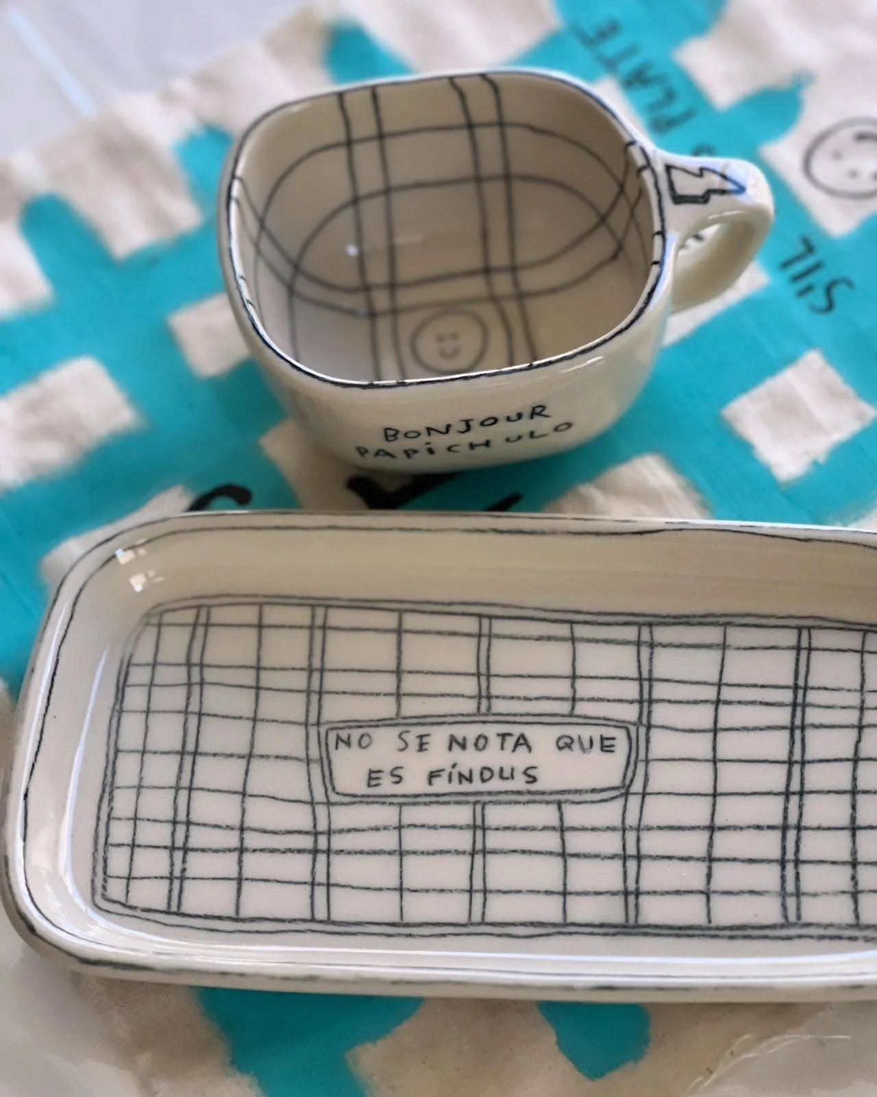 Taza y caja decorativas con frases, con diseño de líneas y cuadros, sobre una superficie con estampado de colores. La taza dice 'Bonjour Pichulo' y la caja tiene la frase 'No se nota que es fin dup'.
