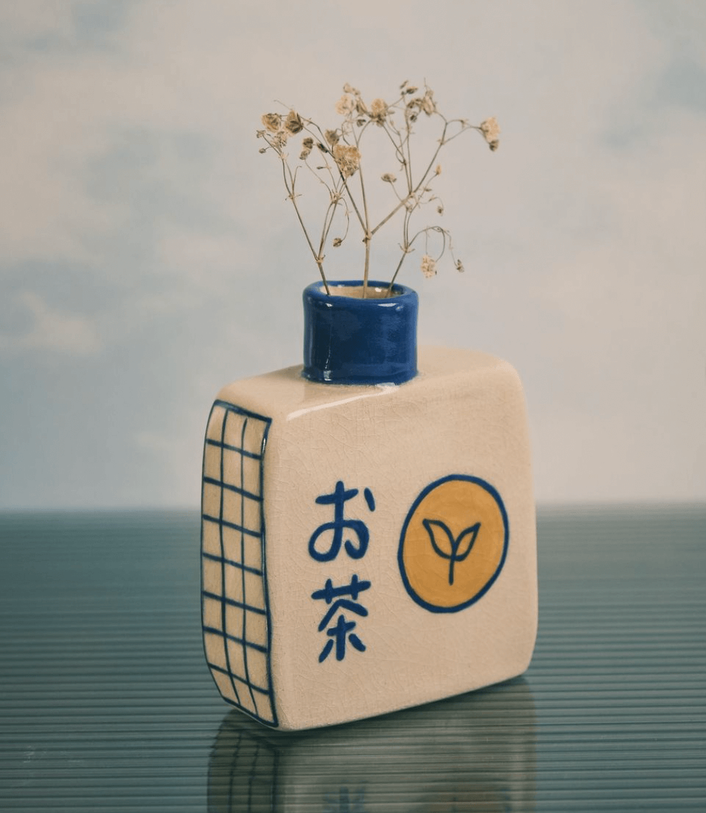 Jarrón azul con flores secas en forma de planta, que se encuentra en una caja o libro decorado con caracteres en japonés y un símbolo de hoja.