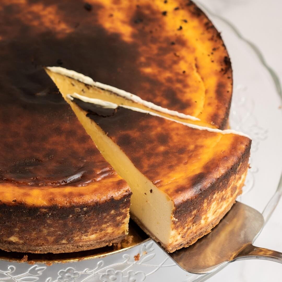 Tarta de queso con costra dorada y corte en una rebanada, sobre un plato de cristal.