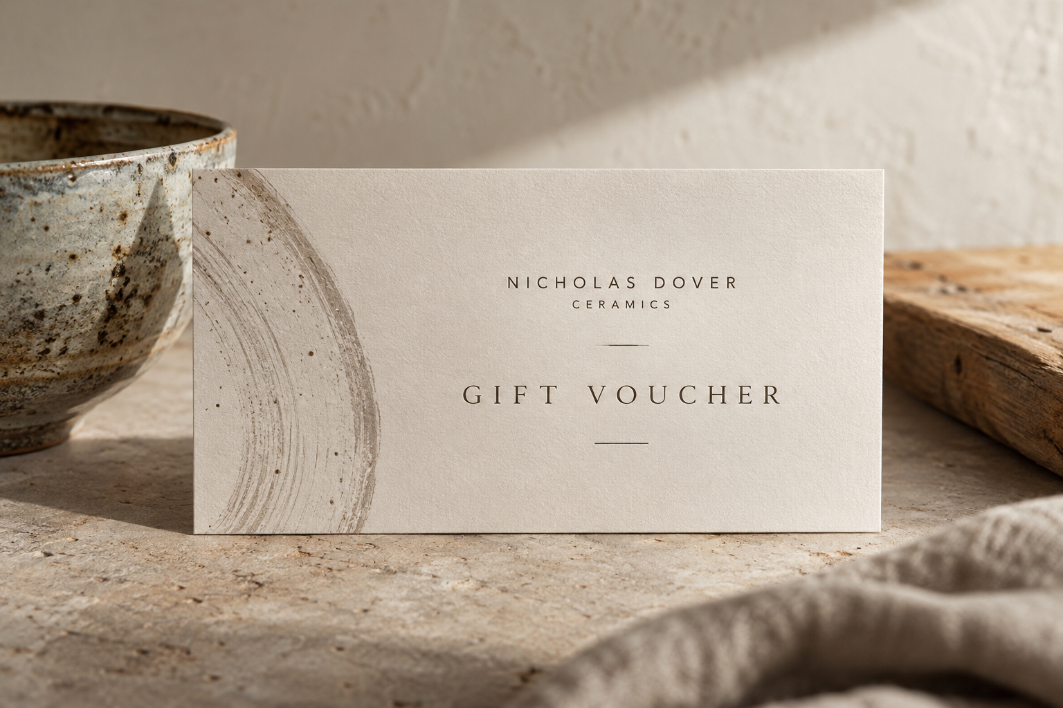 A gift voucher from Nicholas Dover Ceramics with a minimalist design and abstract brushstroke on textured paper, placed on a stone surface next to a rustic bowl and a piece of wood.