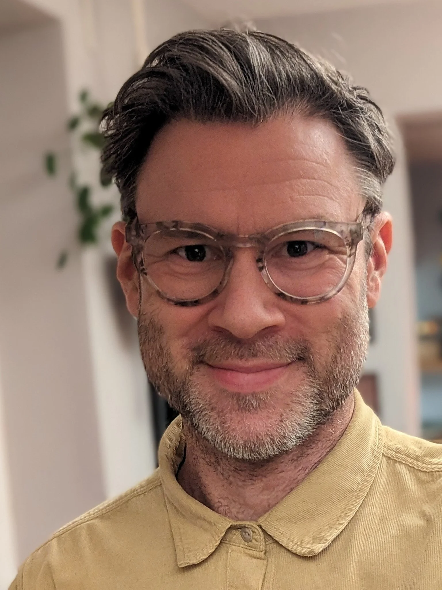 Nicholas Dover with glasses, short gray hair, and facial stubble, wearing a yellow shirt, smiling indoors with a blurred background.