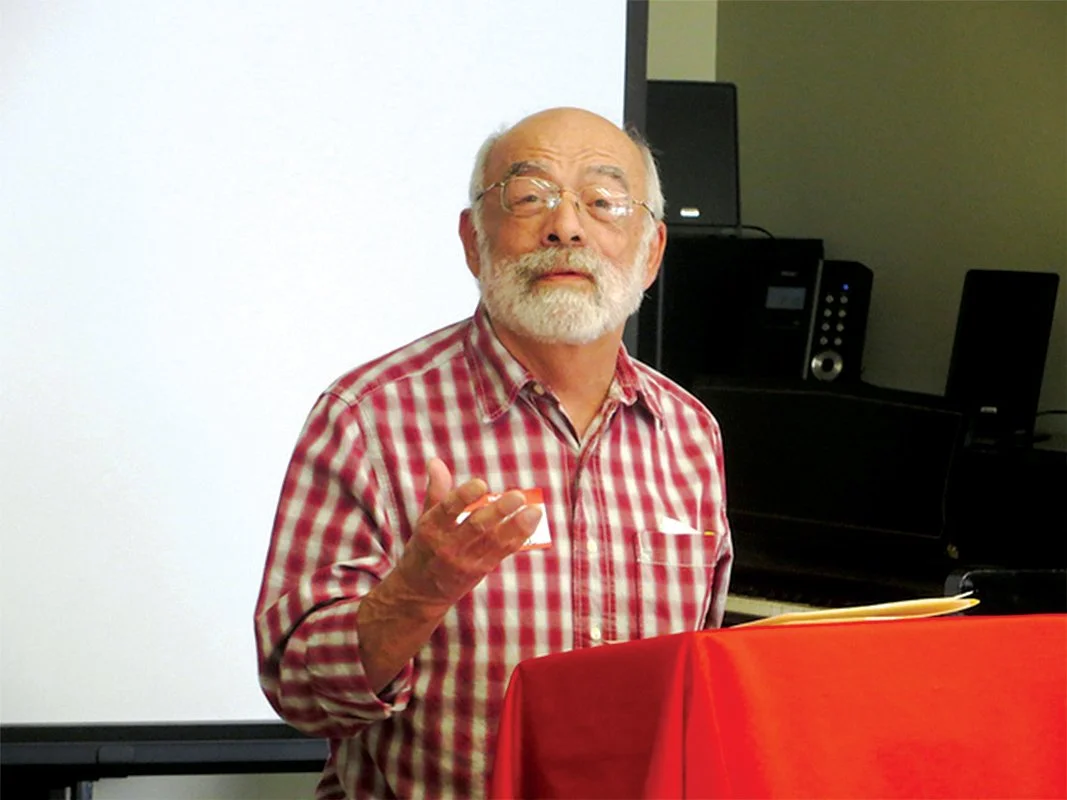 Older male presenter with glasses and a beard standing at a podium, gesturing while addressing an audience in a classroom or meeting room