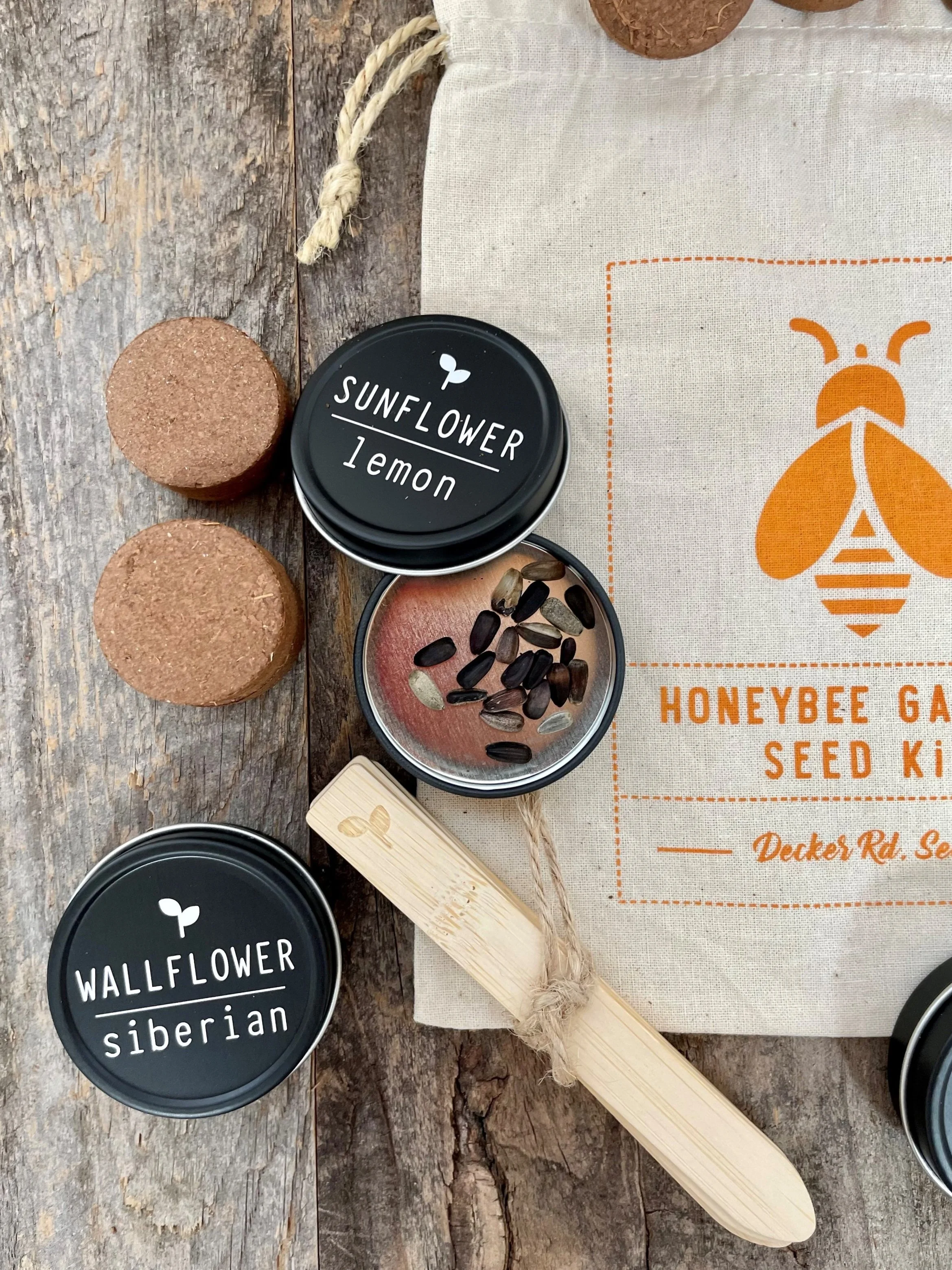Containers of sunflower lemon seeds and wallflower siberian seeds, a seed propagator stick, and a cloth bag with a honeybee garden seed kit logo on a wooden surface.