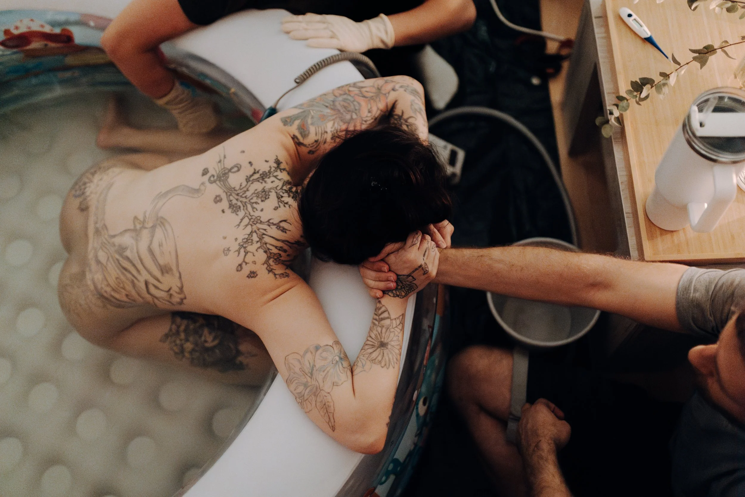 Person mit Tattoo auf dem Rücken liegt in einer Badewanne, während eine andere Person mit ausgezogenen Armen den Kopf hält.