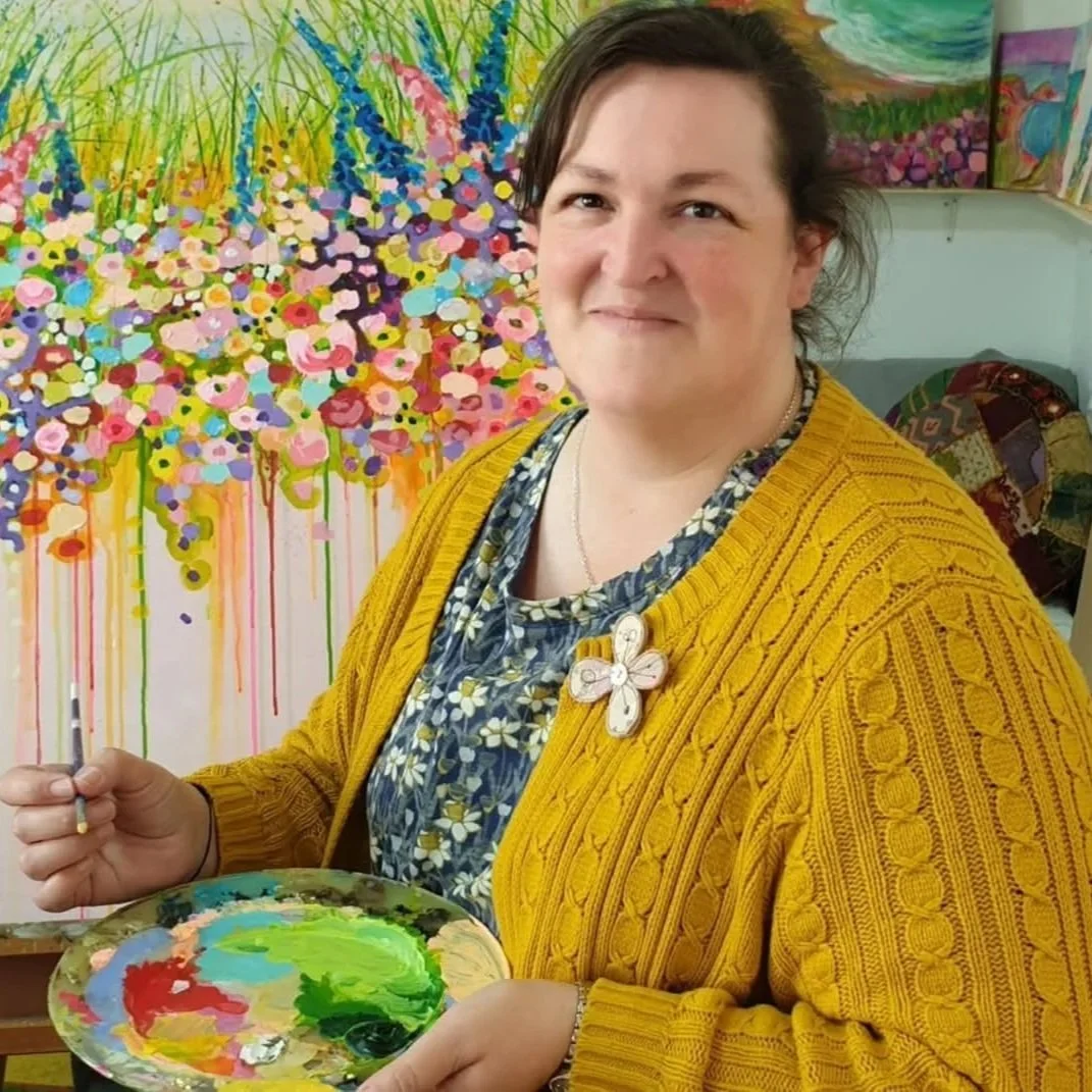 A woman, Jackie Reynolds Artist based in Devon UK, with short dark hair wearing a yellow cardigan and floral dress, holding a paintbrush and palette, standing in front of a colourful floral abstract painting.