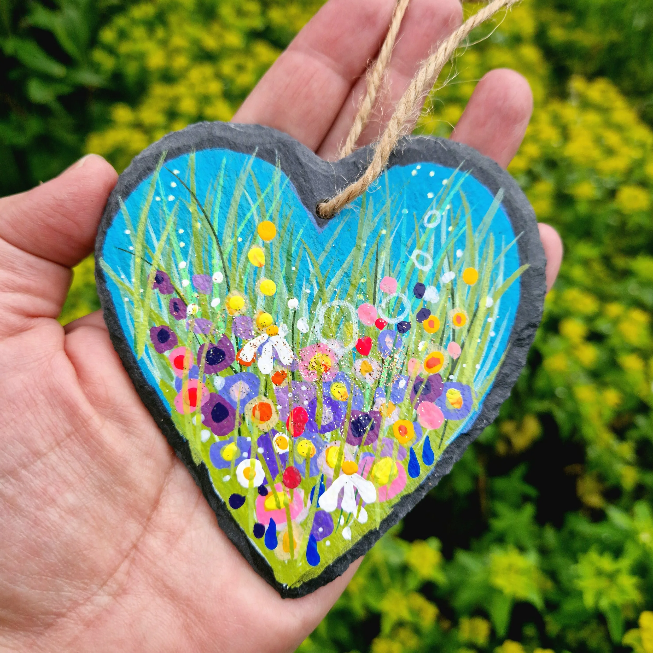 A hand holding a decorated heart-shaped slate hanging decoration with a colourful painted design of flowers and a blue sky, against a blurred green outdoor background.