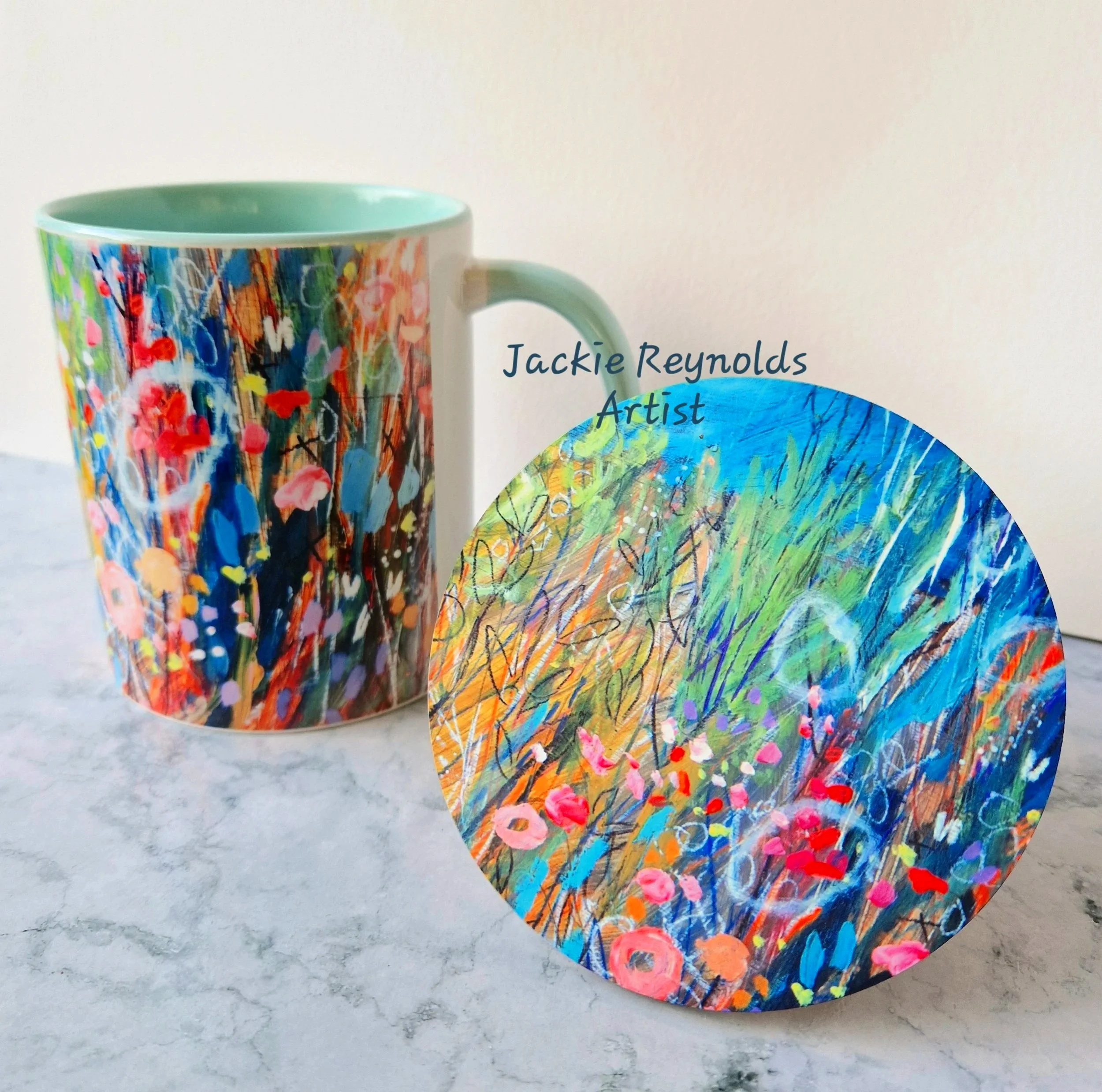 Close-up of a colourful ceramic mug and coaster with abstract floral painted design, placed on a white marble surface.
