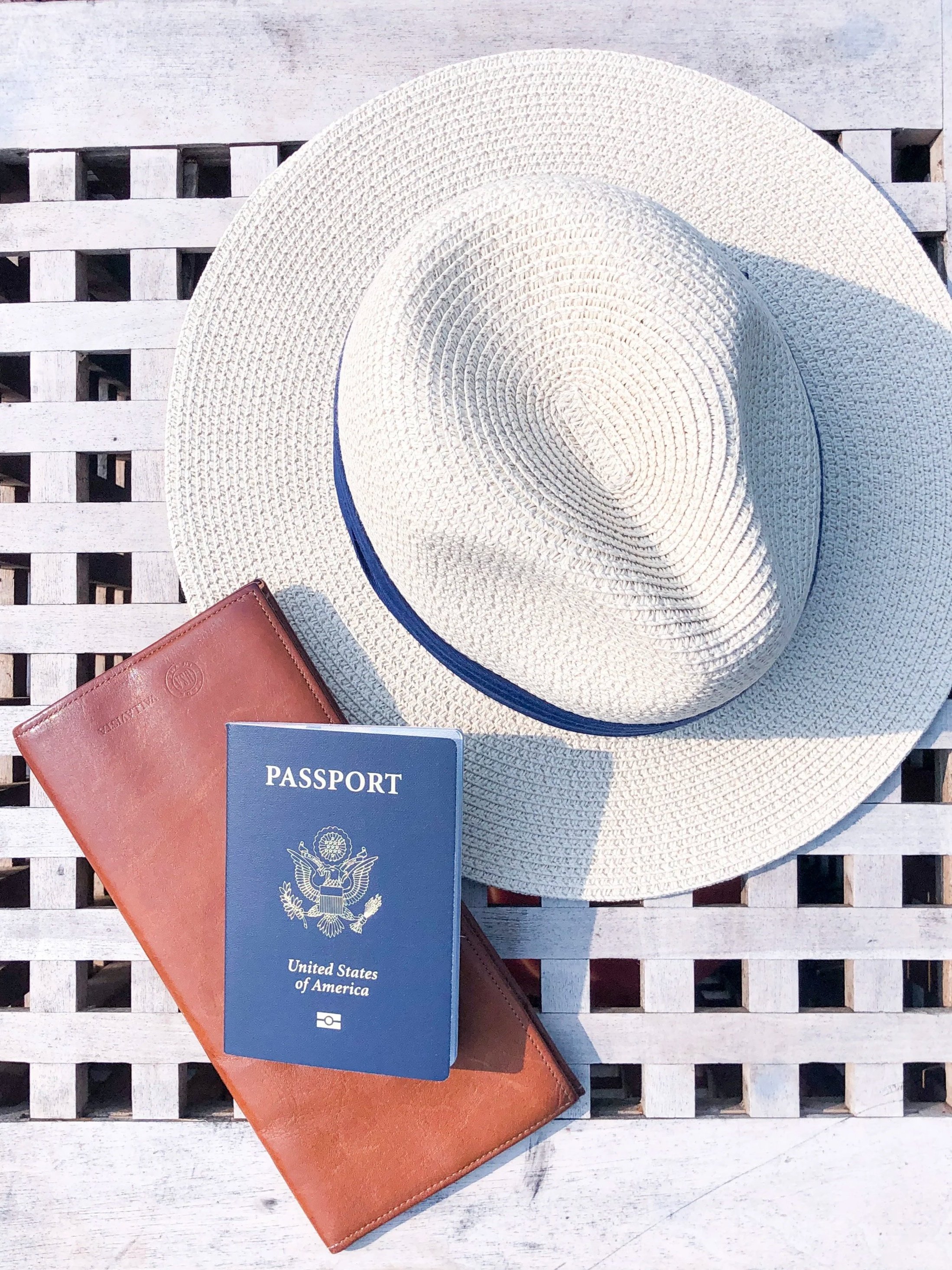 A white woven hat, a United States passport, and a brown wallet arranged on a wooden slatted surface.