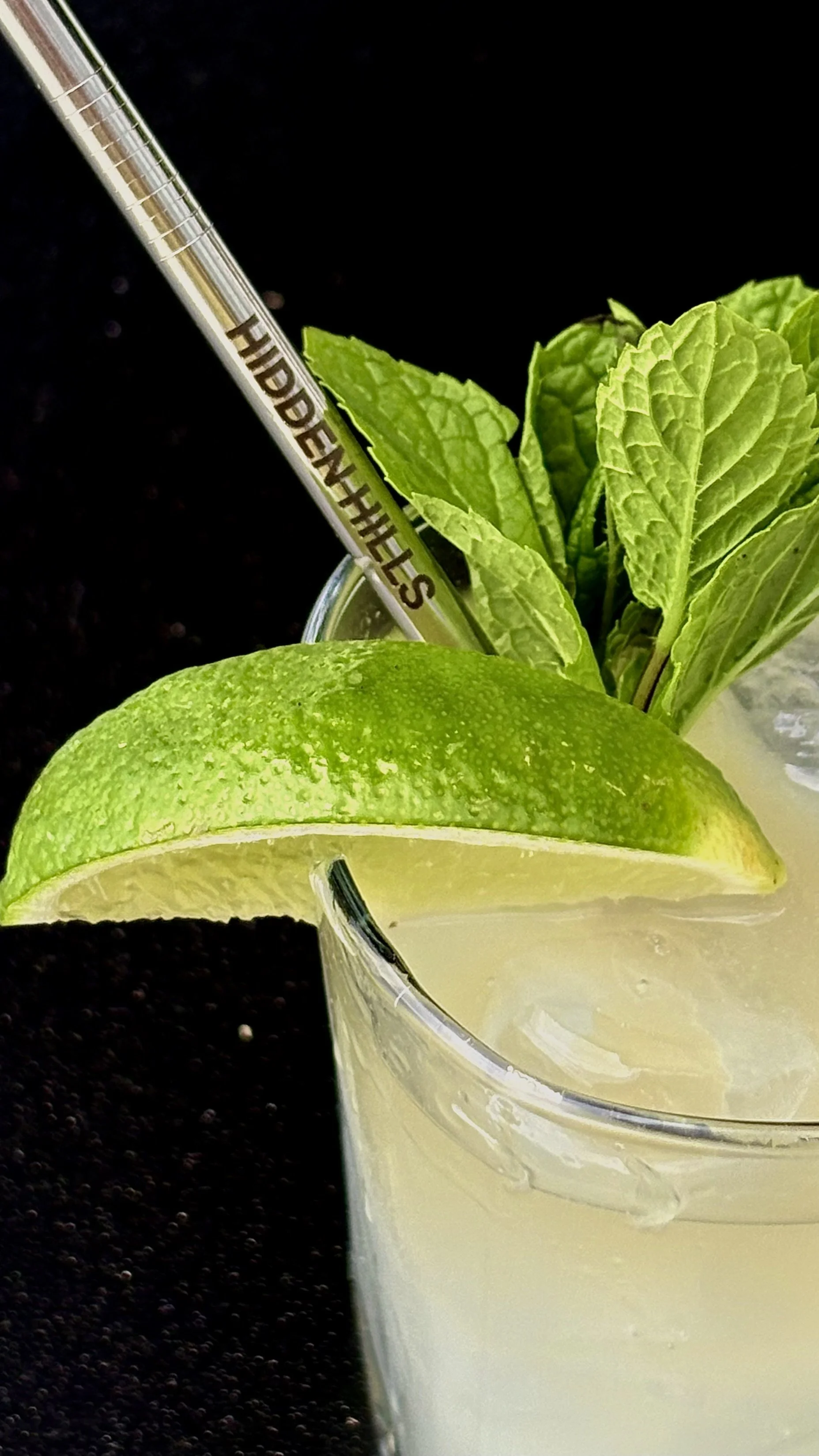A cocktail garnished with a lime wedge and mint leaves, with a metal straw labeled 'HIDDEN WHEELS', in a glass with ice, on a black background.