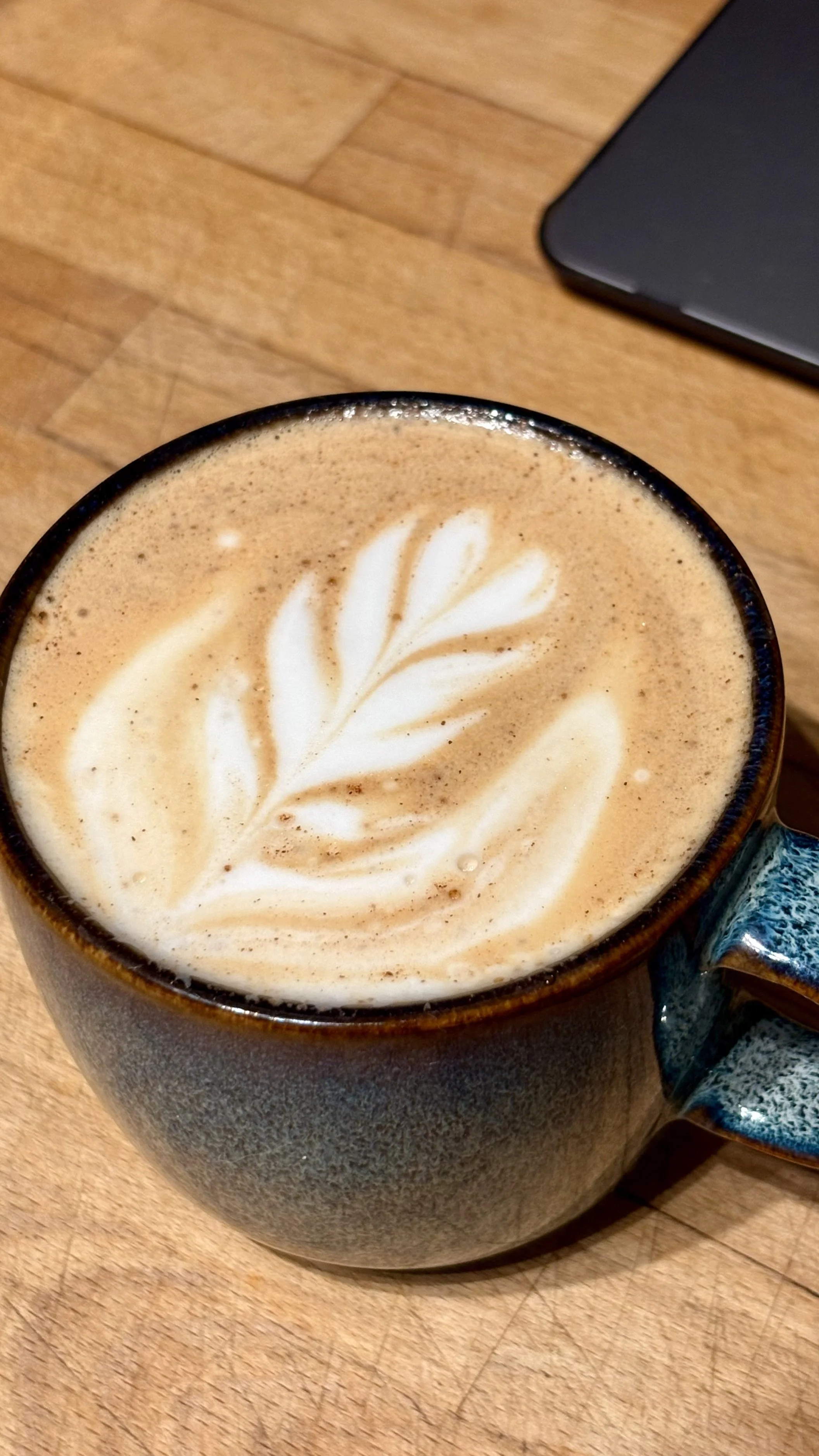 A cup of coffee with latte art, featuring a leaf design, on a wooden table with a laptop partially visible in the background.
