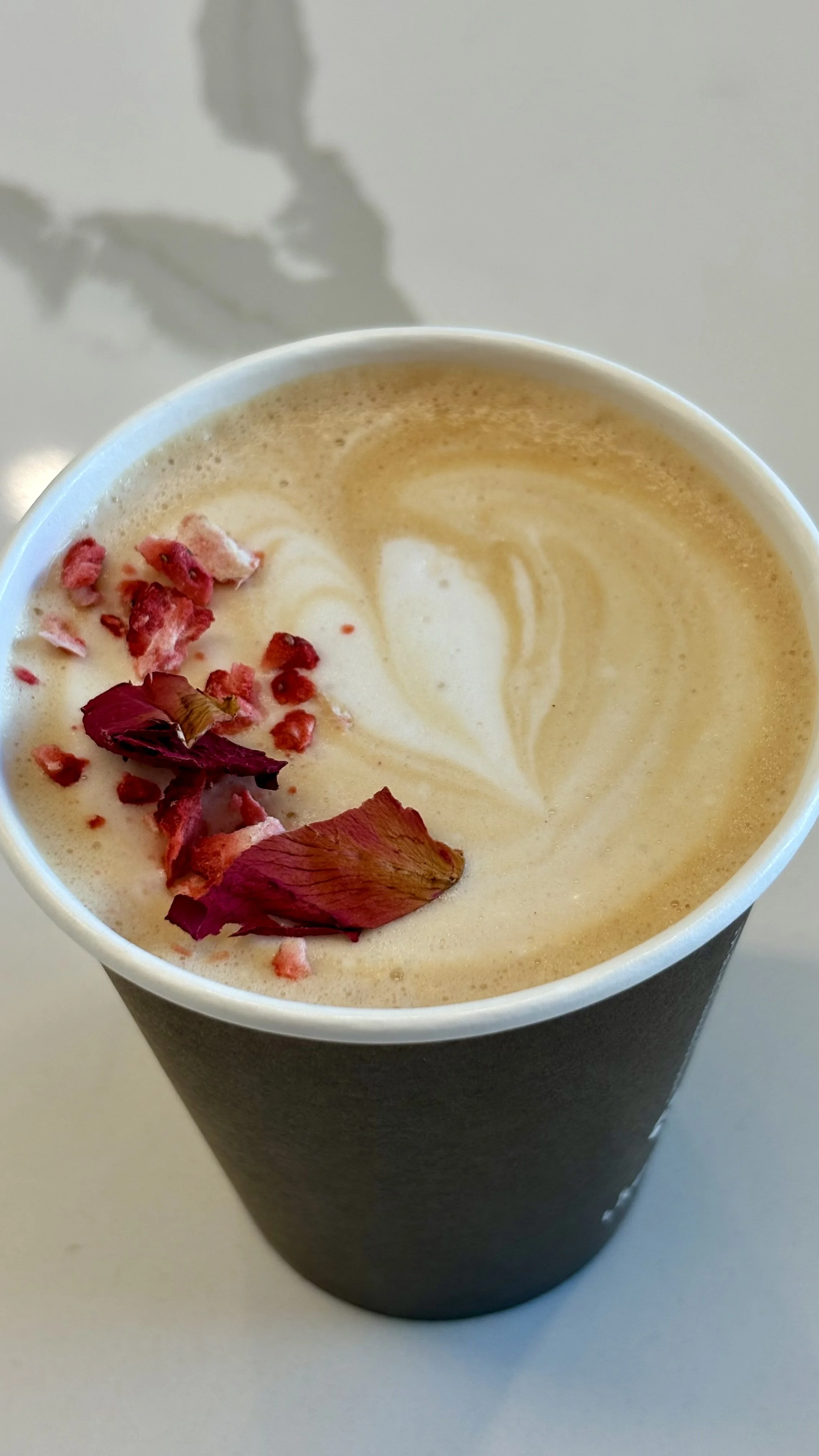 A disposable cup filled with a latte or cappuccino, topped with foam, garnished with rose petals and dried flower pieces.
