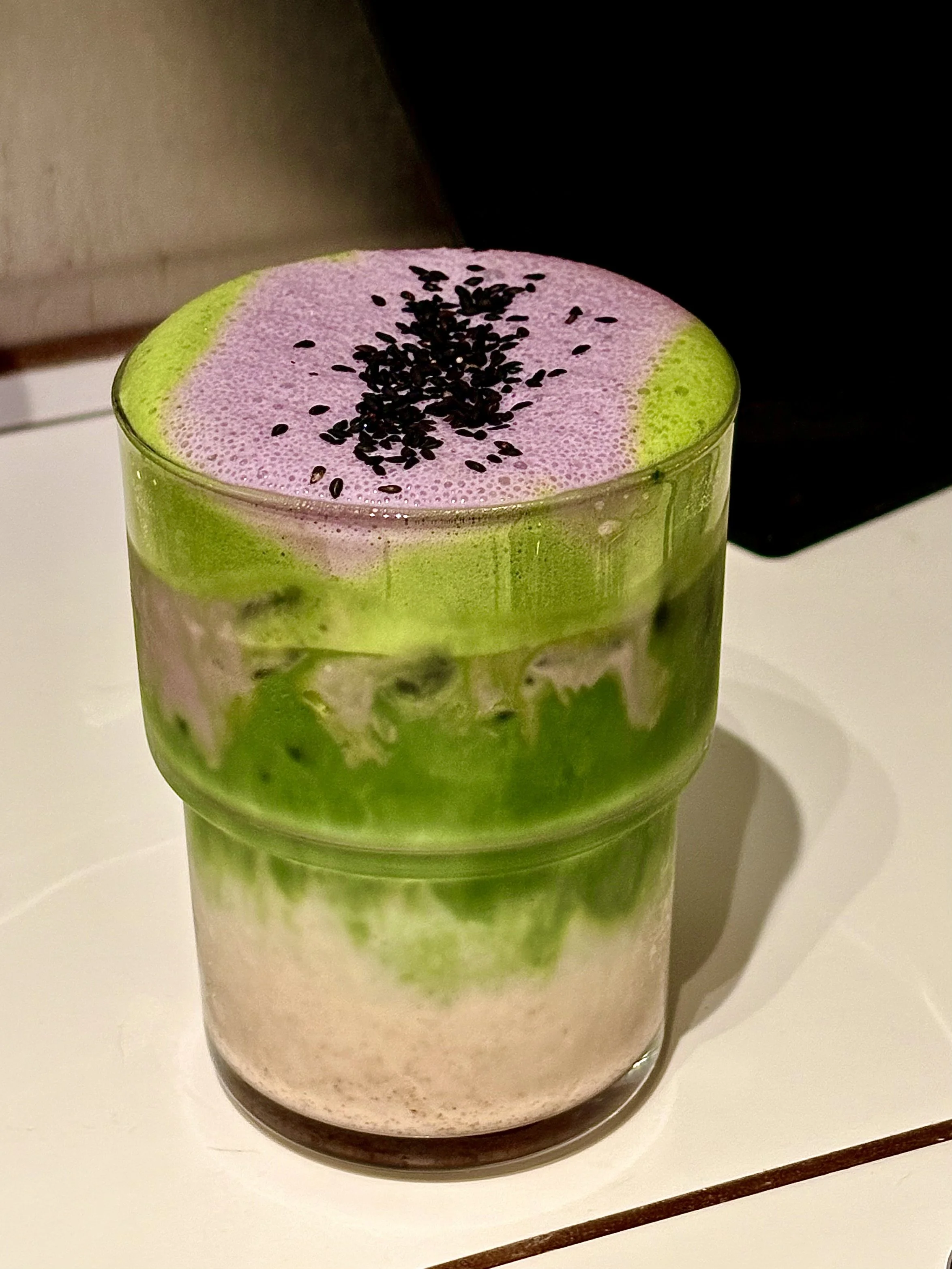 A layered beverage in a tall glass with green, pink, and beige sections, topped with black sesame seeds.