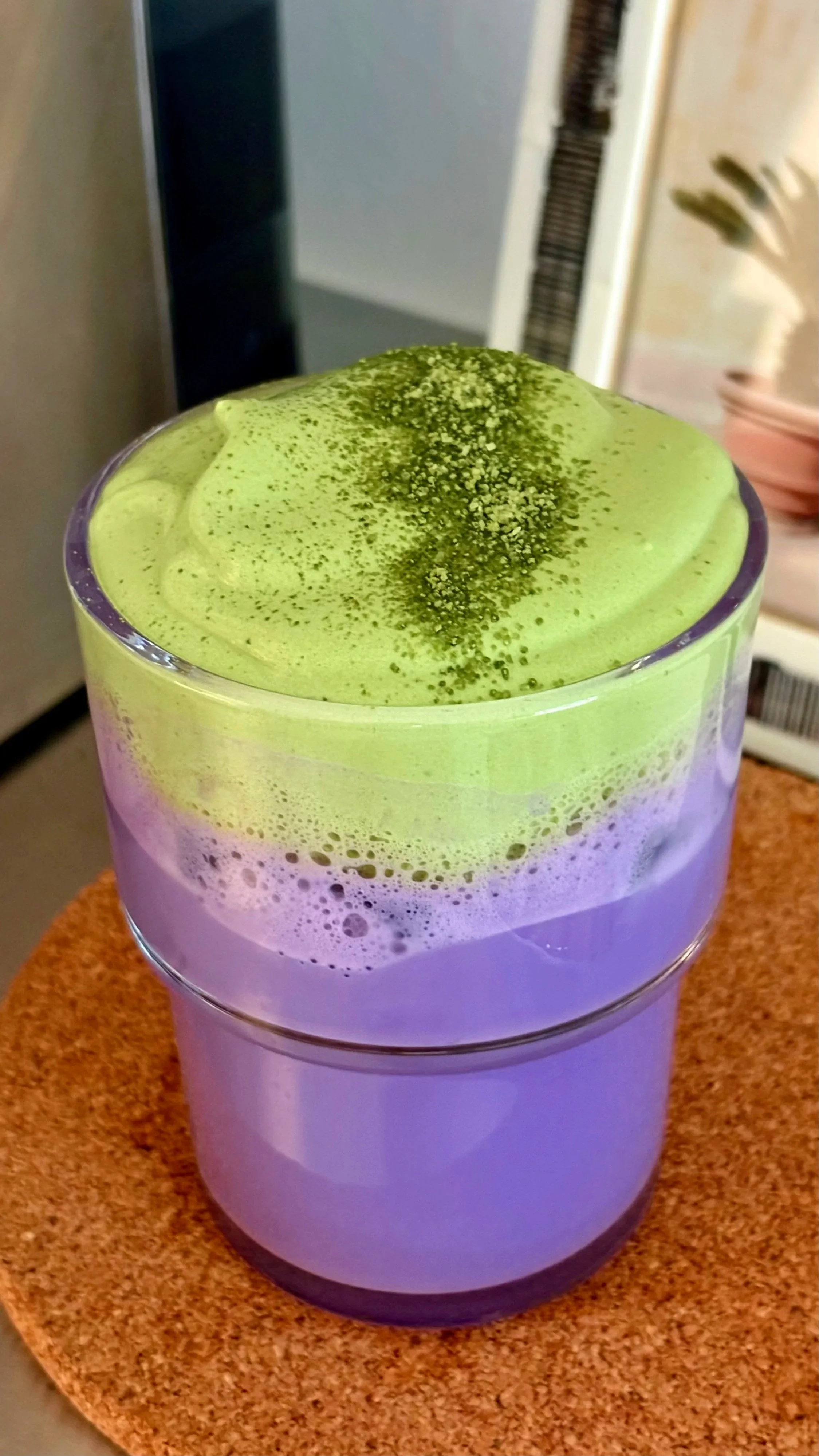 A layered beverage in a clear glass, topped with a green powder, possibly matcha, on green and purple foamy layers, placed on a cork coaster.