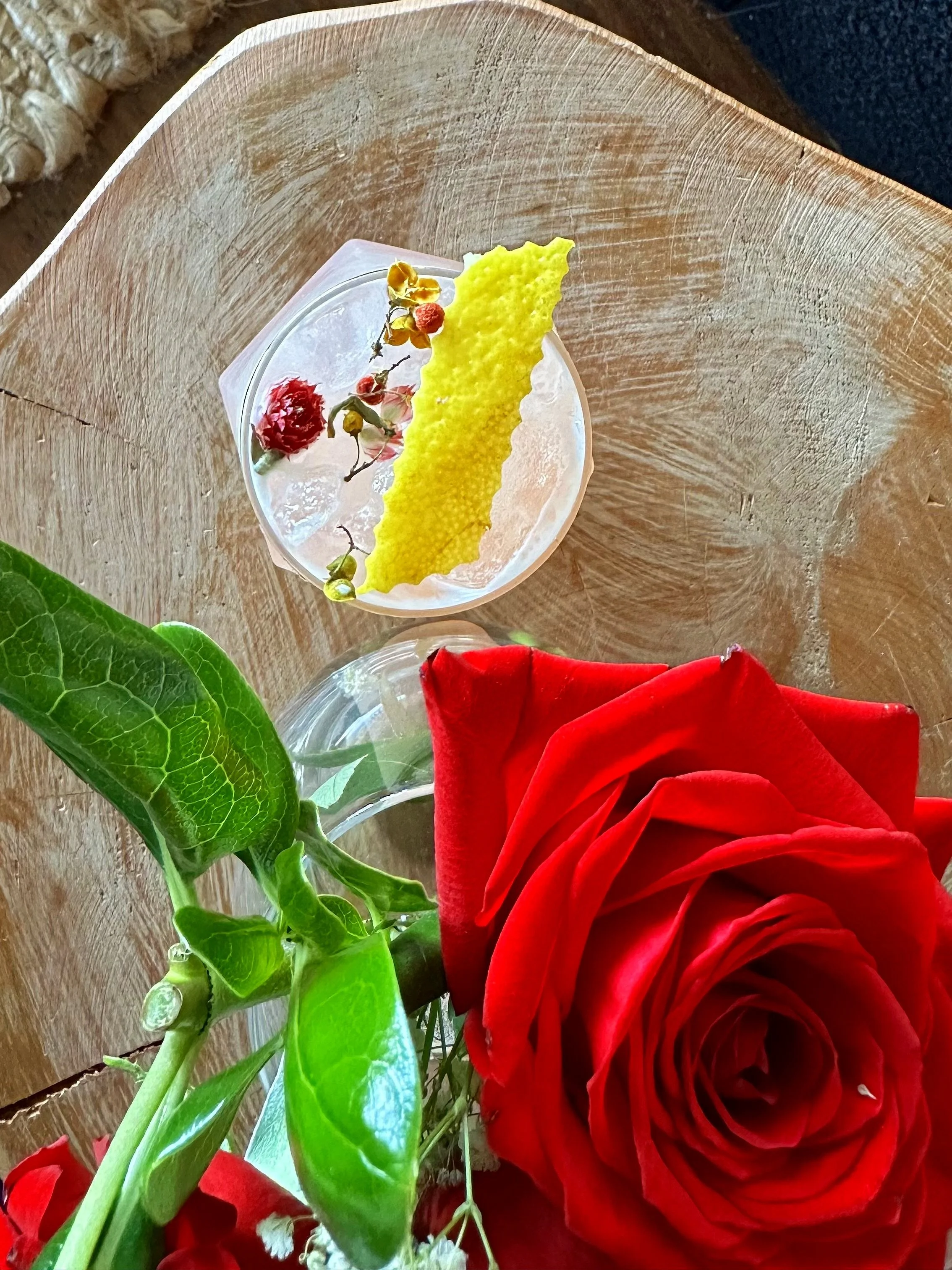 Close-up of red roses with green leaves, a yellow textured object, and small dried flowers on a glass dish, placed on a wooden surface.