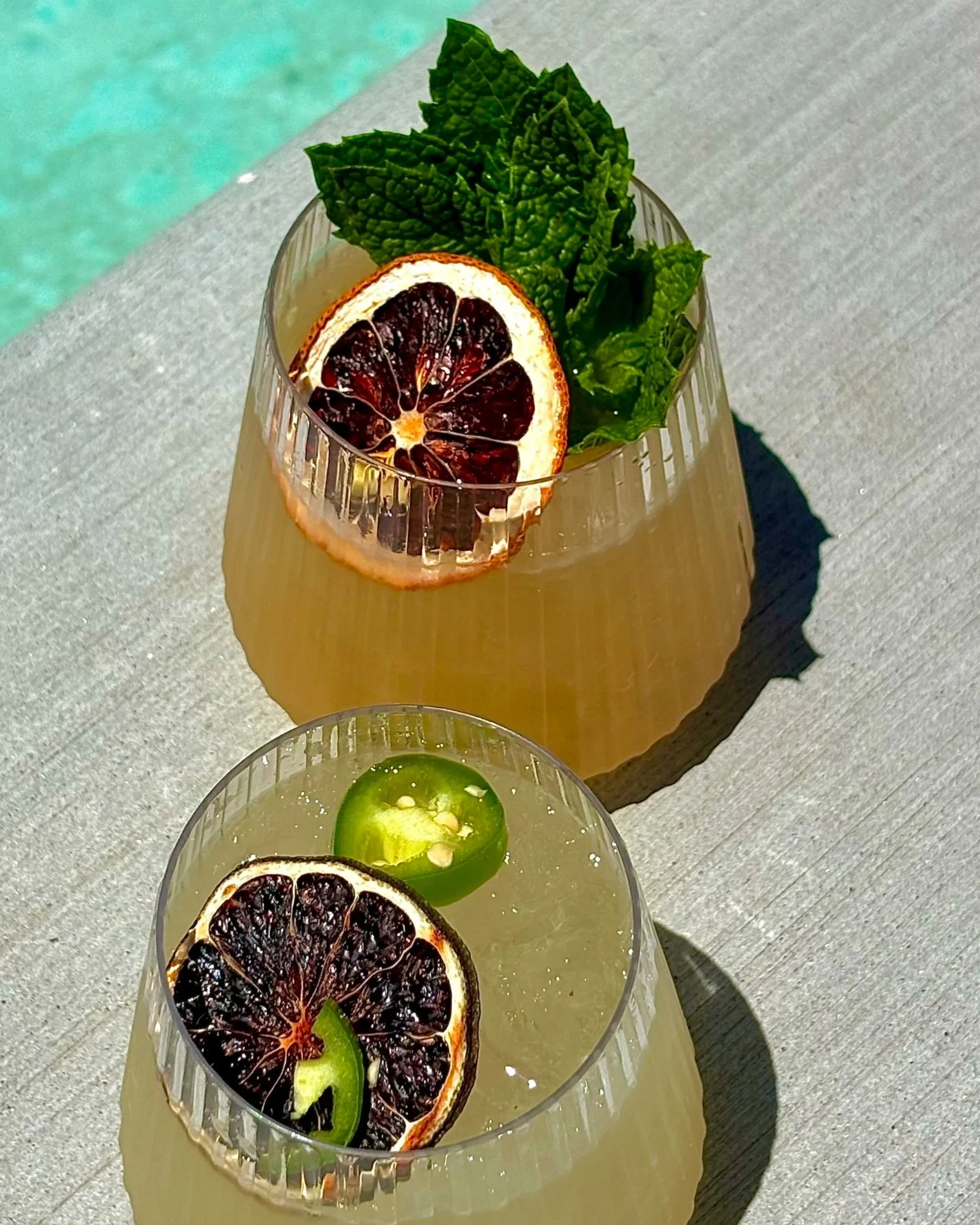 A glass of a yellow-colored drink garnished with a slice of dried orange, a slice of lime, and fresh mint leaves, sitting on a light-colored surface in sunlight.
