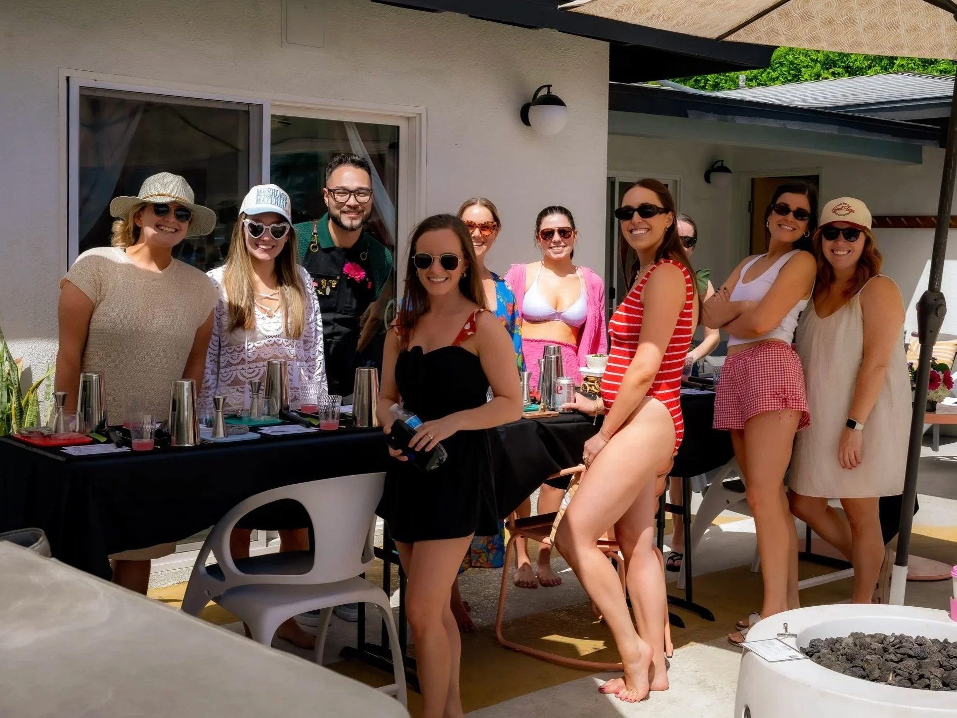 Group of women and one man enjoying a gathering outdoors on a sunny day, standing around a table with drinks and food, some wearing sunglasses, hats, and casual summer clothes.
