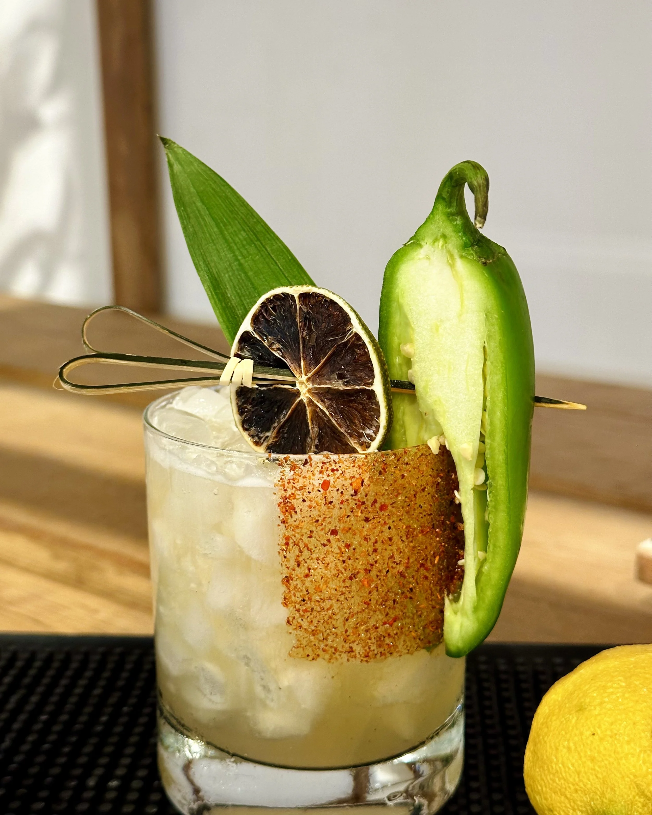 A close-up of a cocktail in a clear glass with a salted rim. Garnished with a lemon wheel, a slice of dried citrus fruit, a green chili Pepper, a green leaf, and a paper umbrella. Part of a lemon is visible on the right side of the image.