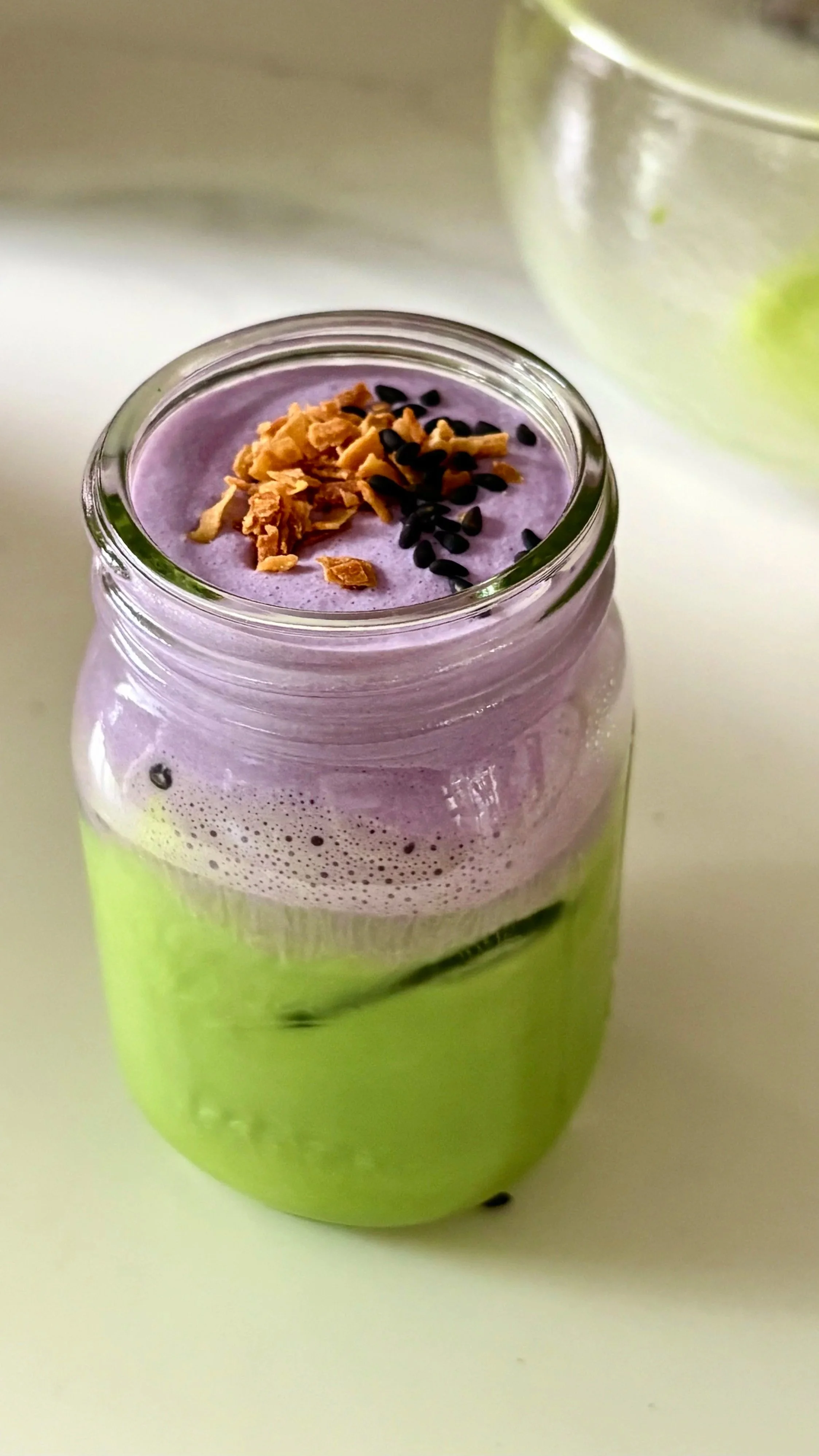 A green smoothie in a mason jar with a purple foam layer on top, garnished with granola and black sesame seeds.