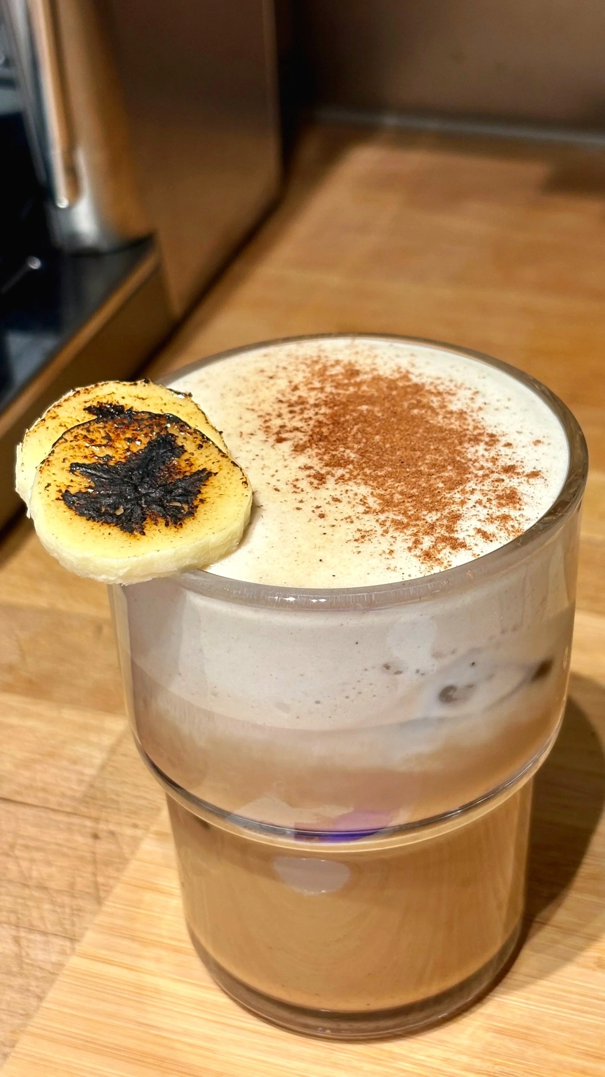 A layered coffee drink in a clear glass topped with a dollop of whipped cream, a dusting of cocoa powder, and a grilled banana slice garnishing the rim.