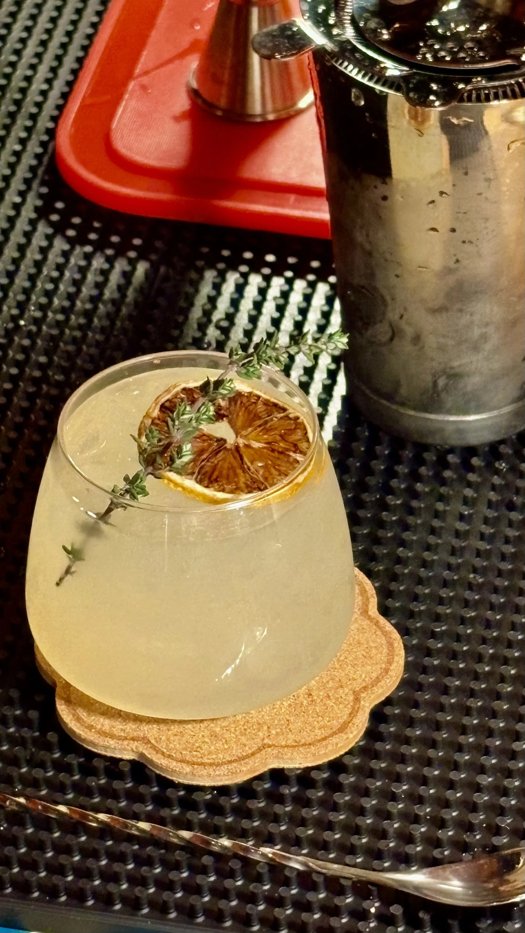 A cocktail glass with a lemon slice and a sprig of thyme on top, placed on a cork coaster on a black textured surface, with a metal grater, a red cutting board, and a metal squeezer in the background.