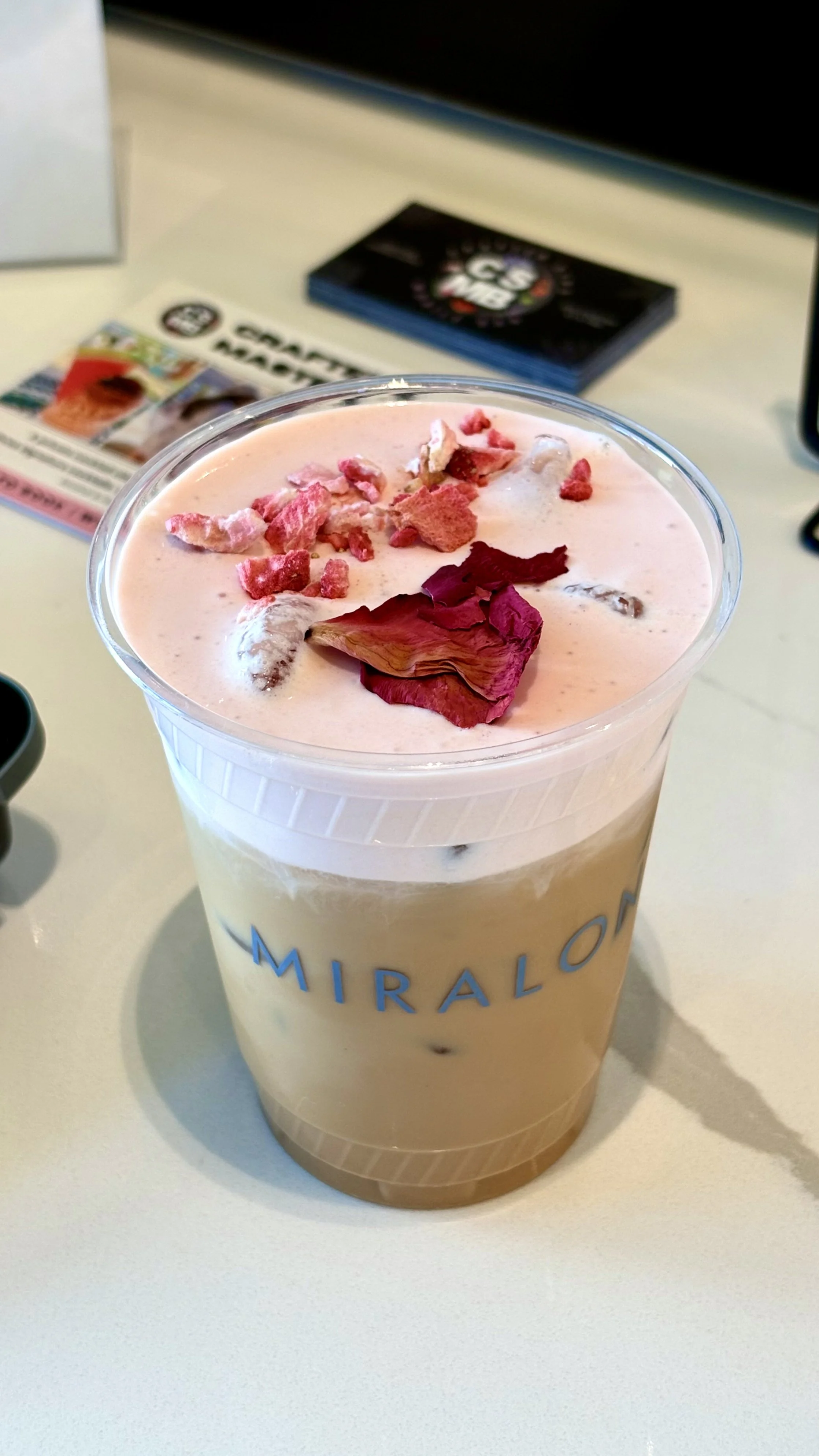 Frosty pink milkshake with rose petals and crushed strawberries on top in a clear cup at Miralo shop, blurred magazines and booklets in the background.