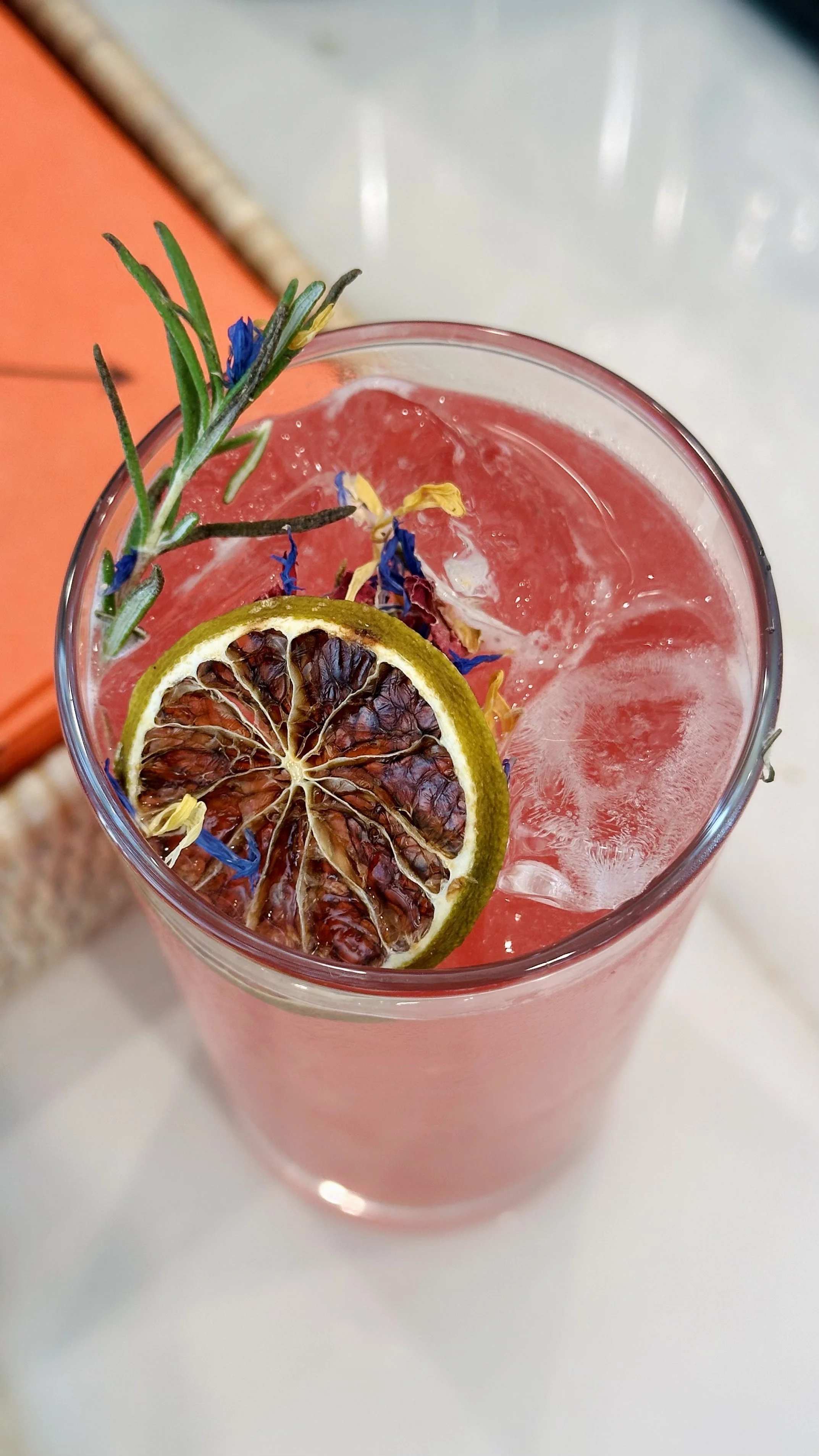 A pink beverage garnished with a dried lemon slice, a sprig of rosemary, and colorful flower petals in a clear glass.