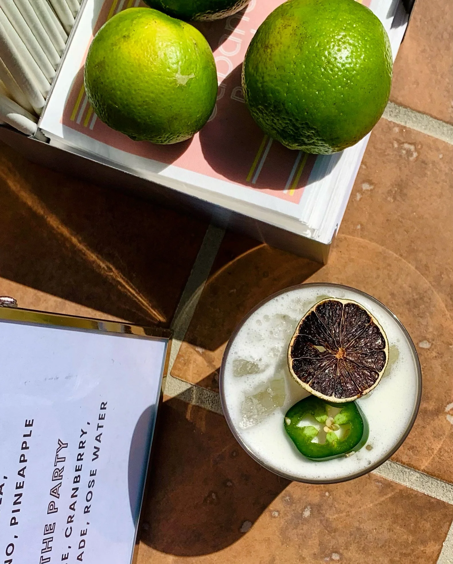 Two fresh green limes on a paper surface, with a dried half lime or lemon and lime slices on a cup filled with a creamy beverage, possibly a cocktail.