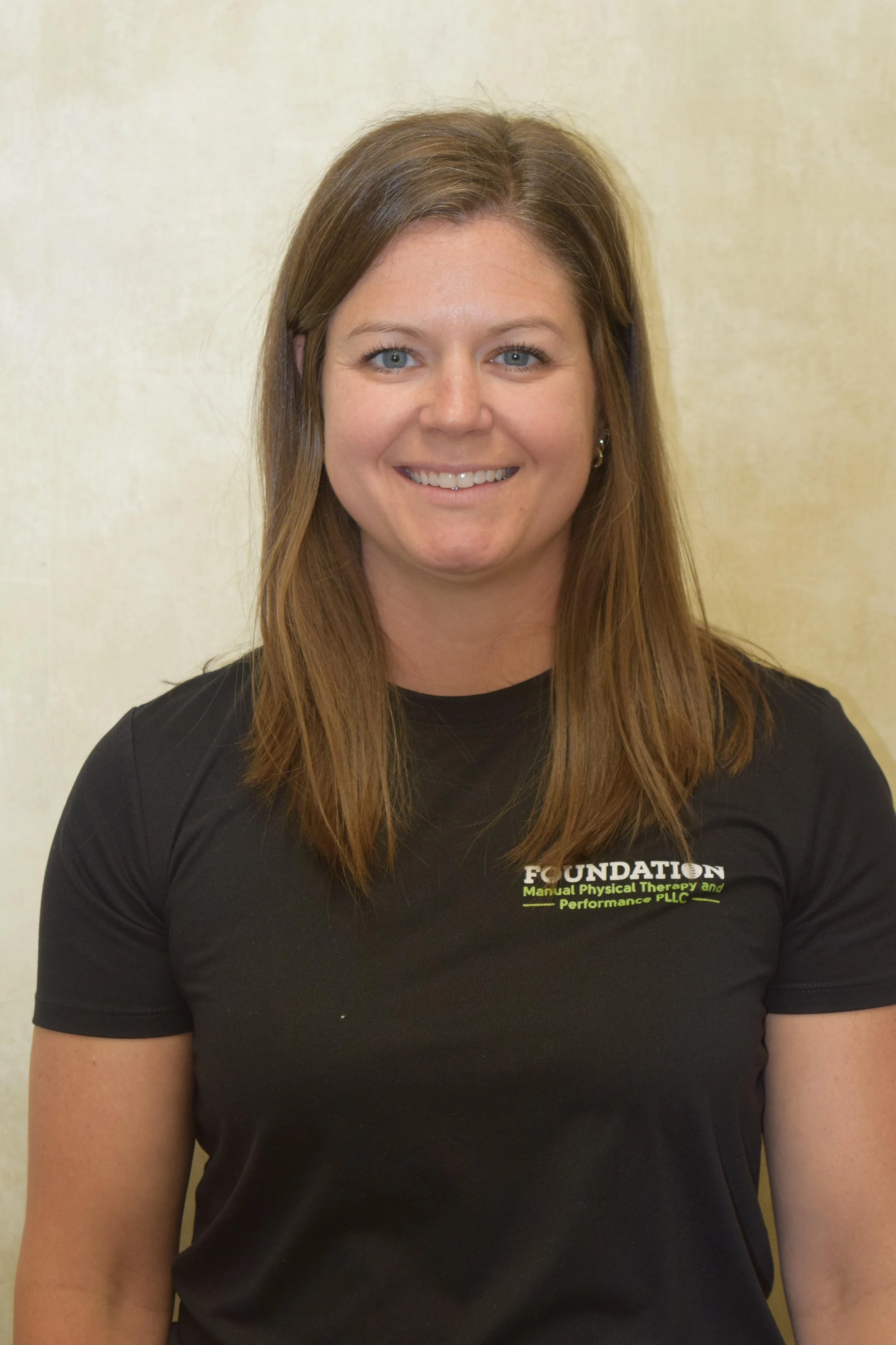 A young woman with long, light brown hair standing against a plain beige wall, smiling, wearing a black T-shirt with green and white text that reads 'FOUNDATION' and some additional smaller text.