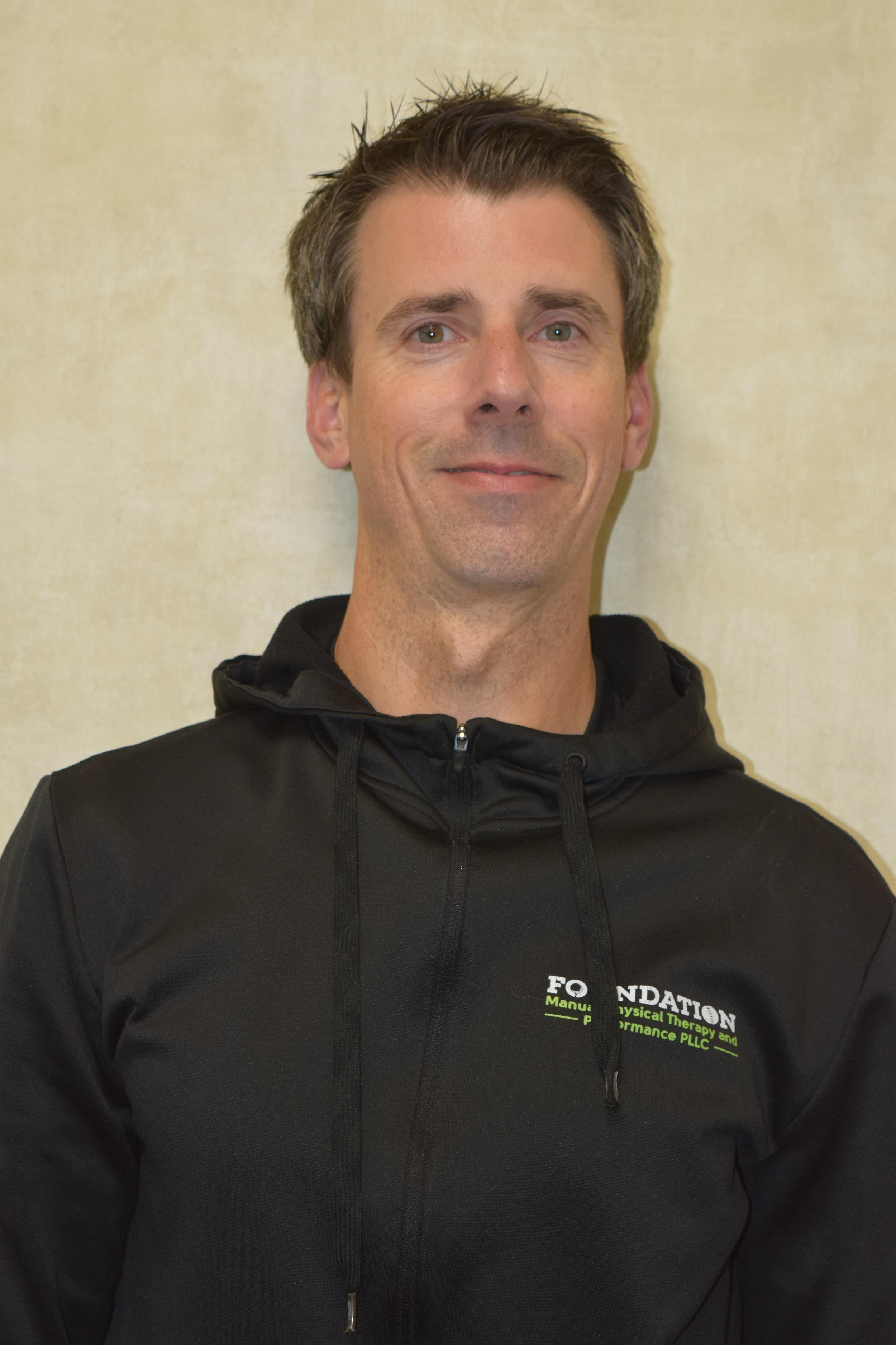 A man with short brown hair and a light beard, wearing a black zip-up jacket with a logo that reads 'FOUNDATION' on the chest, standing against a plain beige wall.