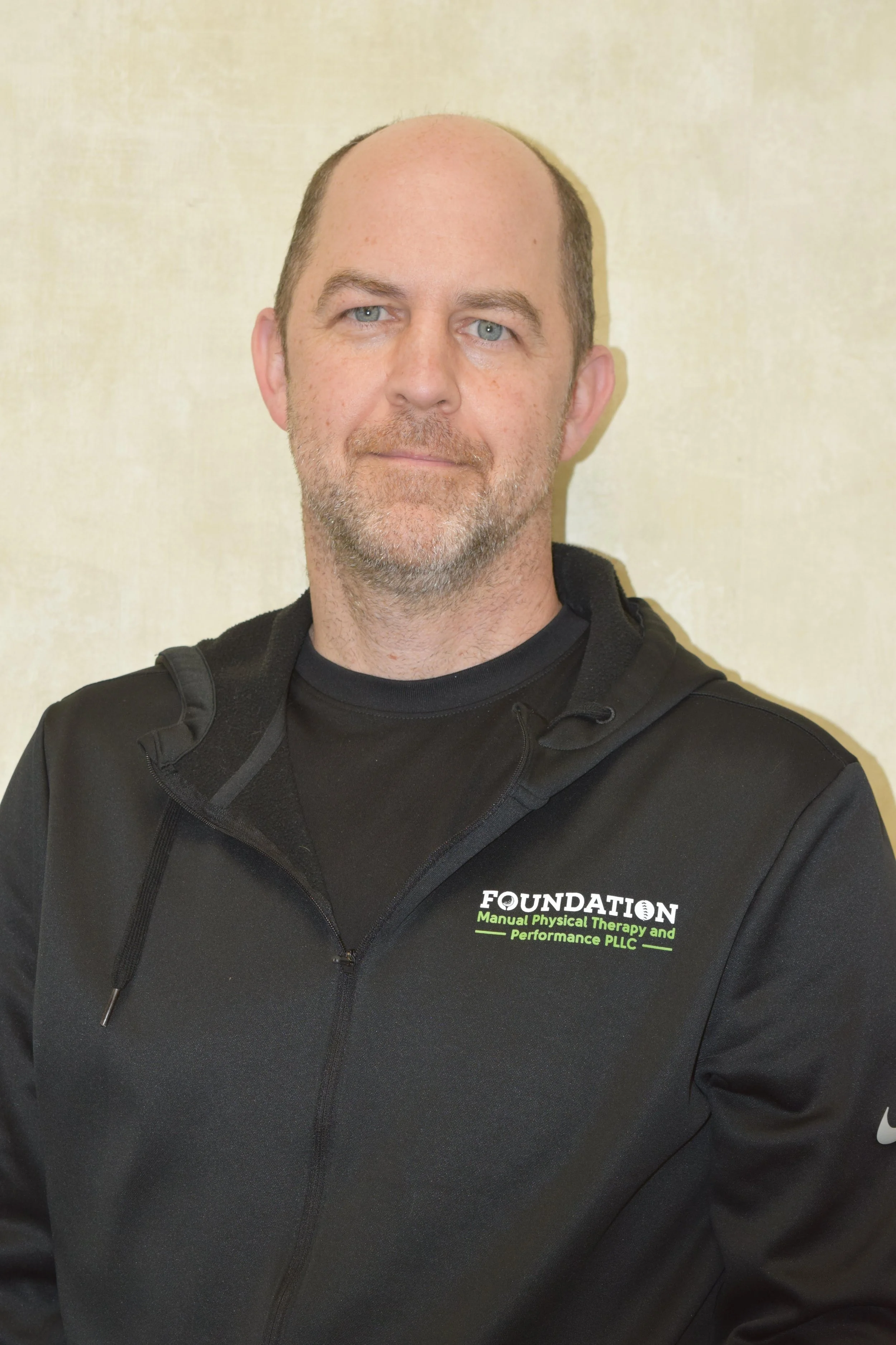 A middle-aged man with a shaved head and light beard, wearing a black athletic jacket over a black T-shirt, standing in front of a plain beige wall, smiling slightly.