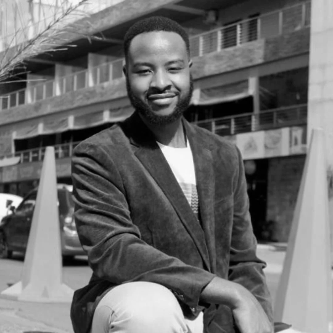 A smiling man with a beard and short hair, wearing a blazer over a T-shirt, sitting outdoors in front of a building with balconies, with cars and traffic cones in the background.