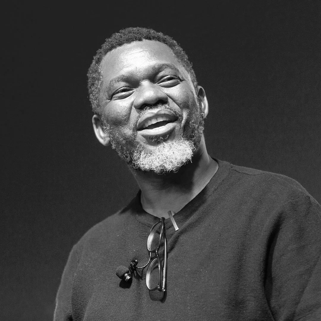 A smiling Black man with a beard and glasses hanging from his shirt, wearing a black shirt, against a dark background.