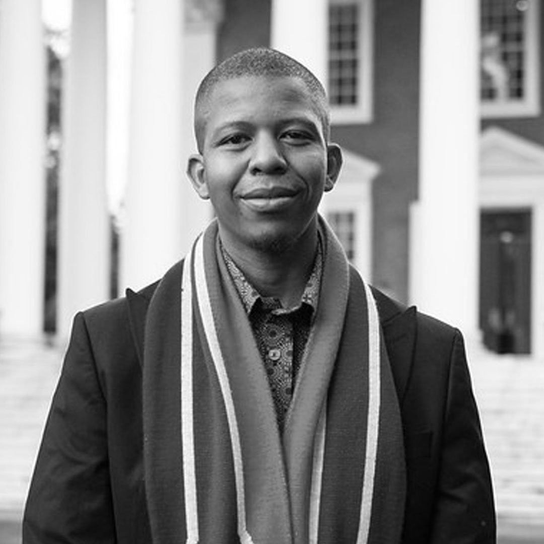 A young man standing outdoors in front of a building with columns, smiling softly at the camera, dressed in a suit with a patterned shirt and a scarf.