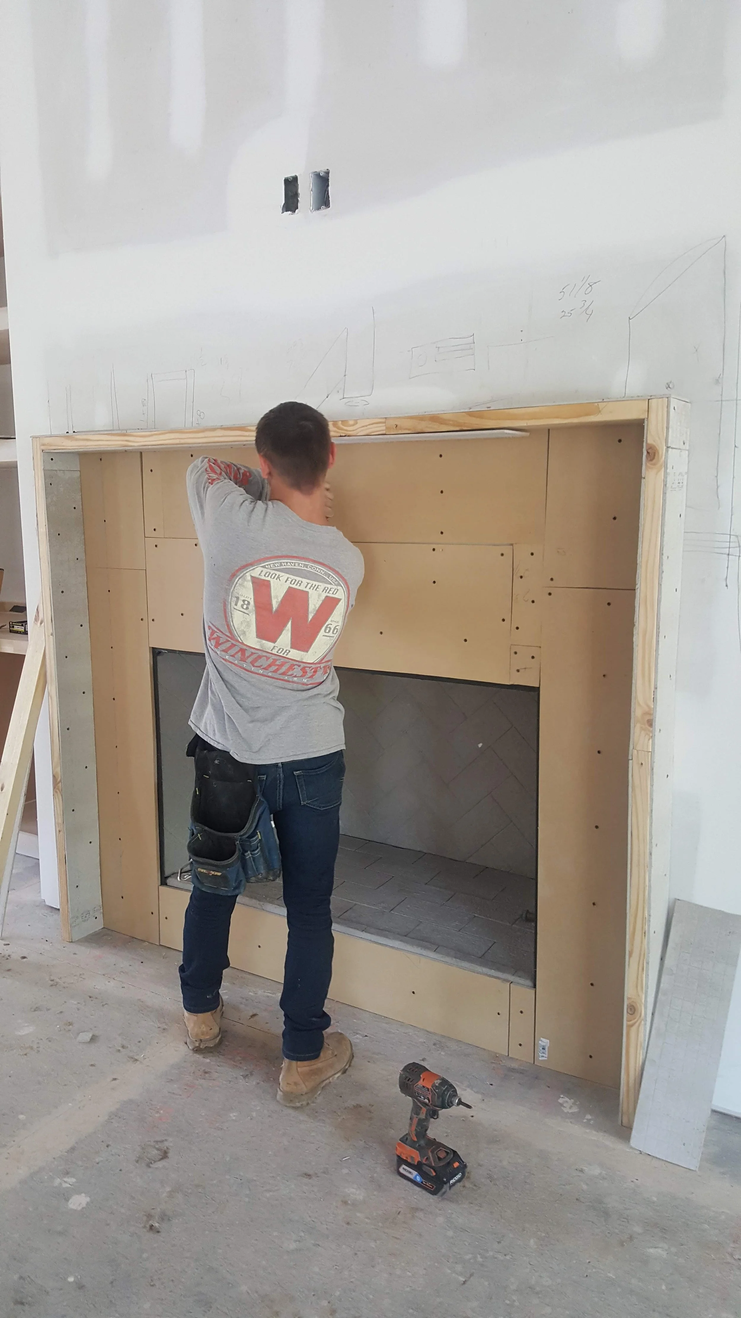 A construction worker standing in front of a partially built fireplace, working on installing or constructing it. There is a power drill on the floor nearby and some sketches and measurements on the wall above.