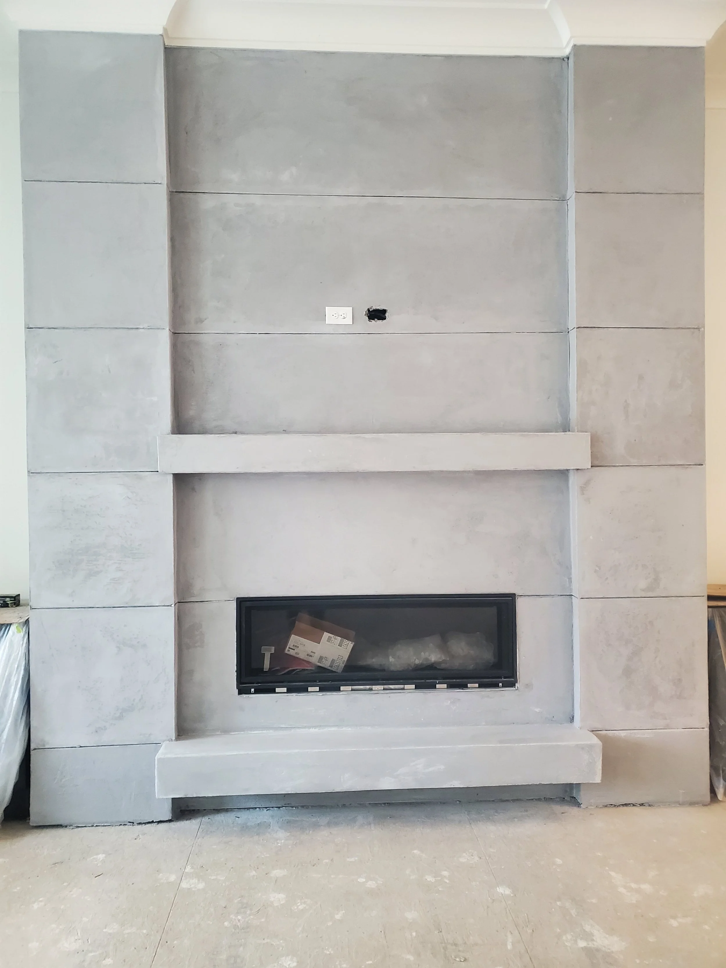 Unfinished gray concrete fireplace with an opening for a firebox and an empty space above, with electrical outlets and a hole for wiring.