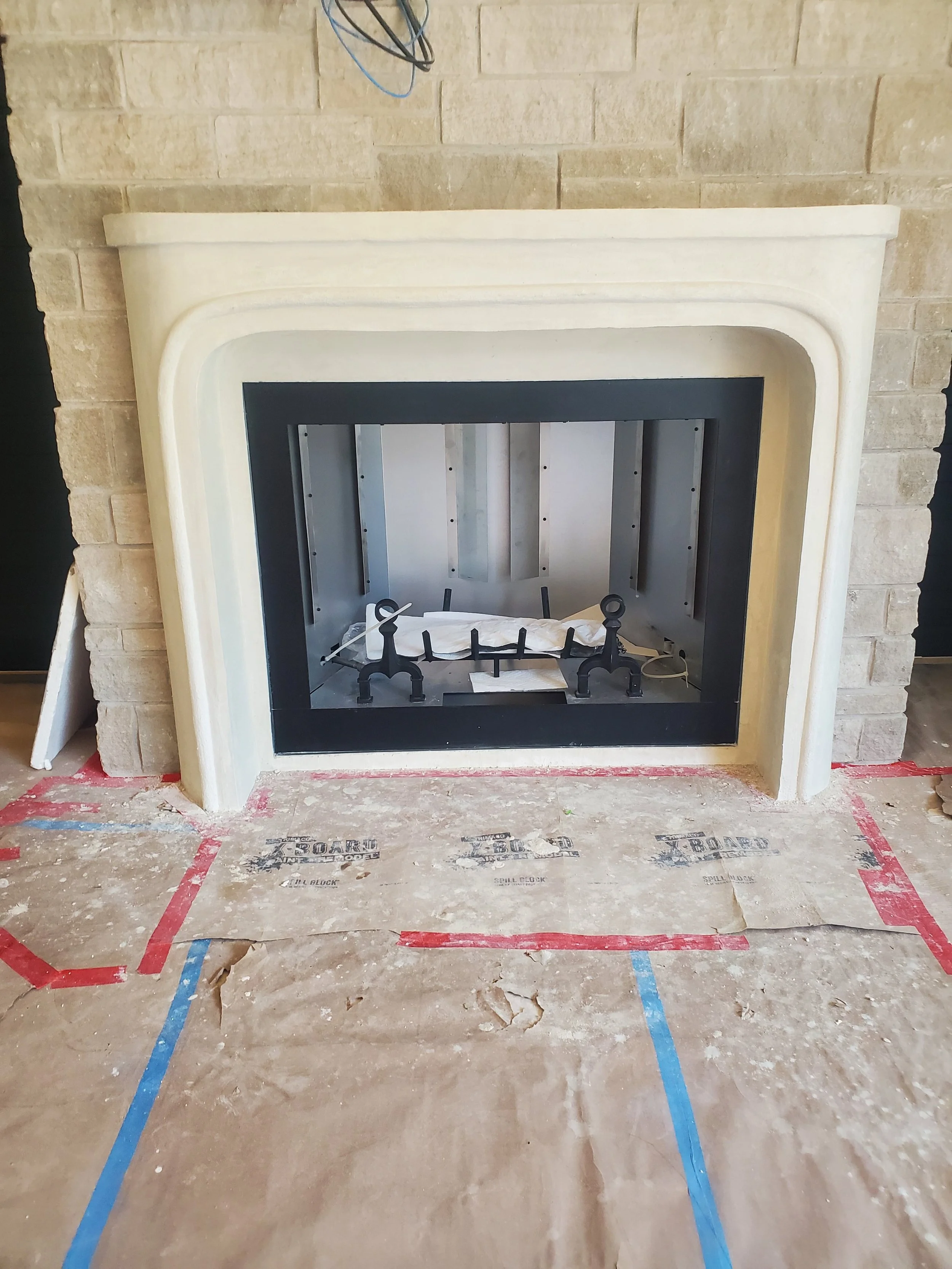 Unfinished fireplace with a cream-colored mantel and firebox, surrounded by beige brick wall and construction floor paper with red and blue tape.