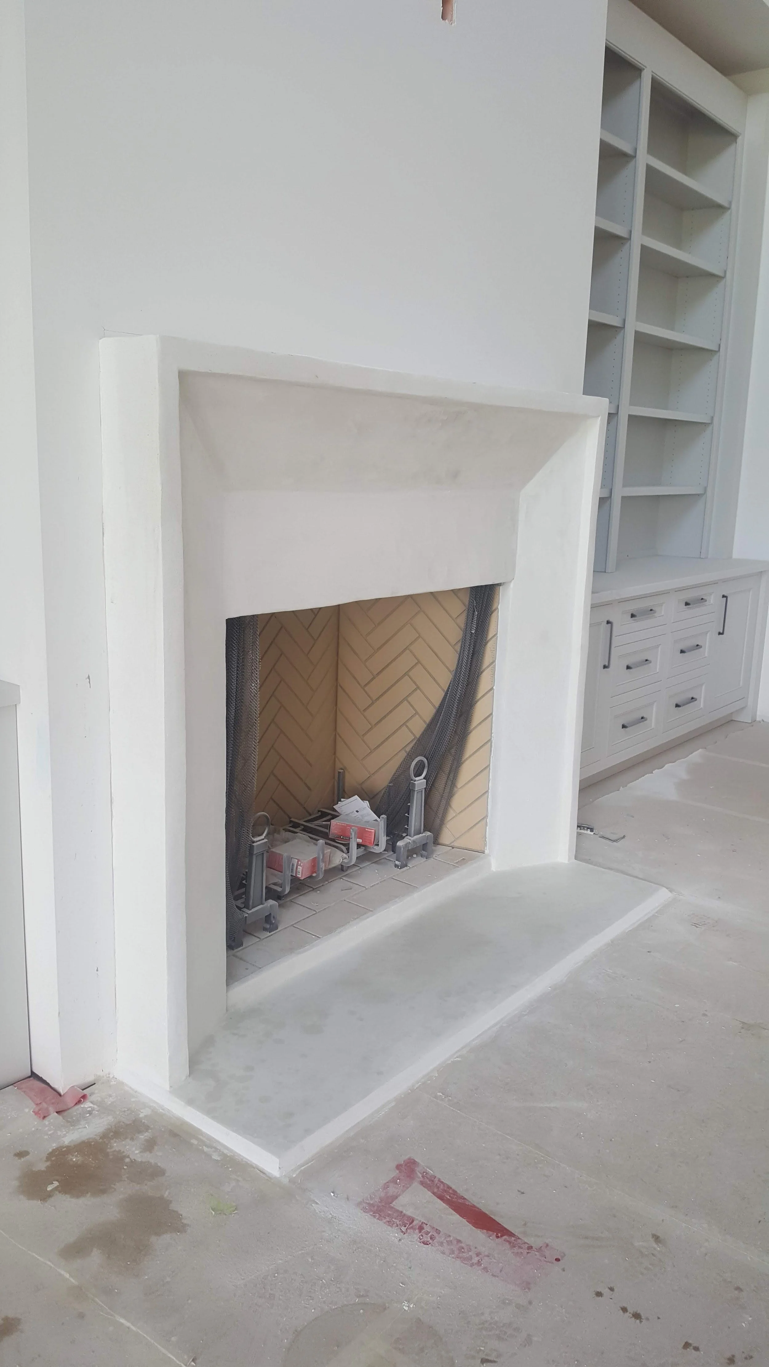 Under construction white fireplace with an unfinished hearth in a residential room. There are building materials and tools visible inside the fireplace opening, with shelves and drawers in the background.