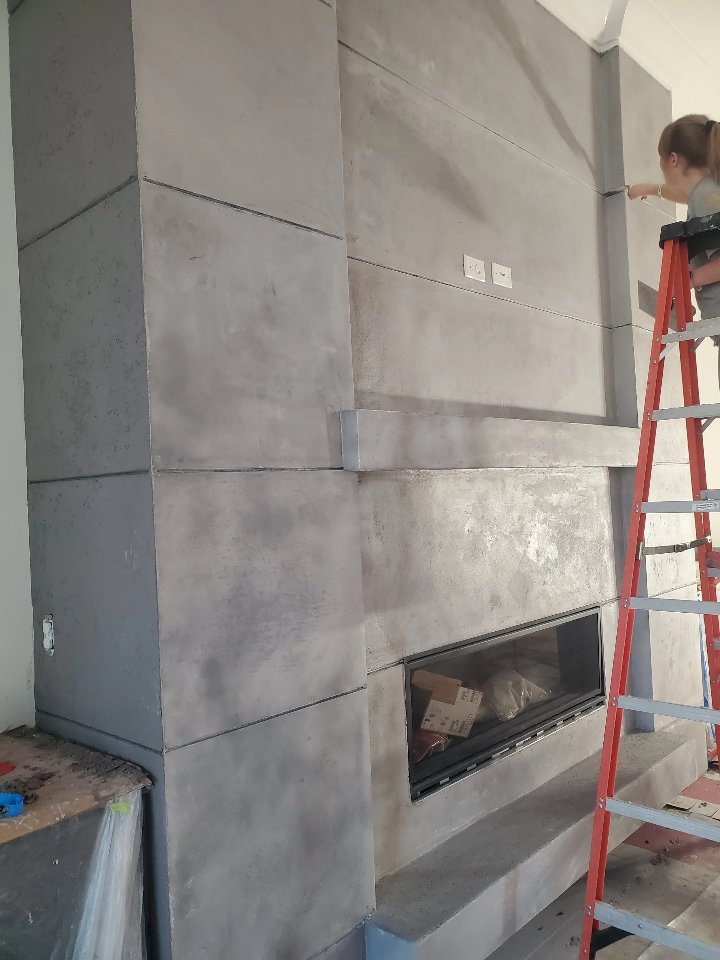 Construction worker on a red ladder installing or inspecting a wall-mounted electrical outlet or switch on a modern concrete wall with a fireplace opening below, in an unfinished room.