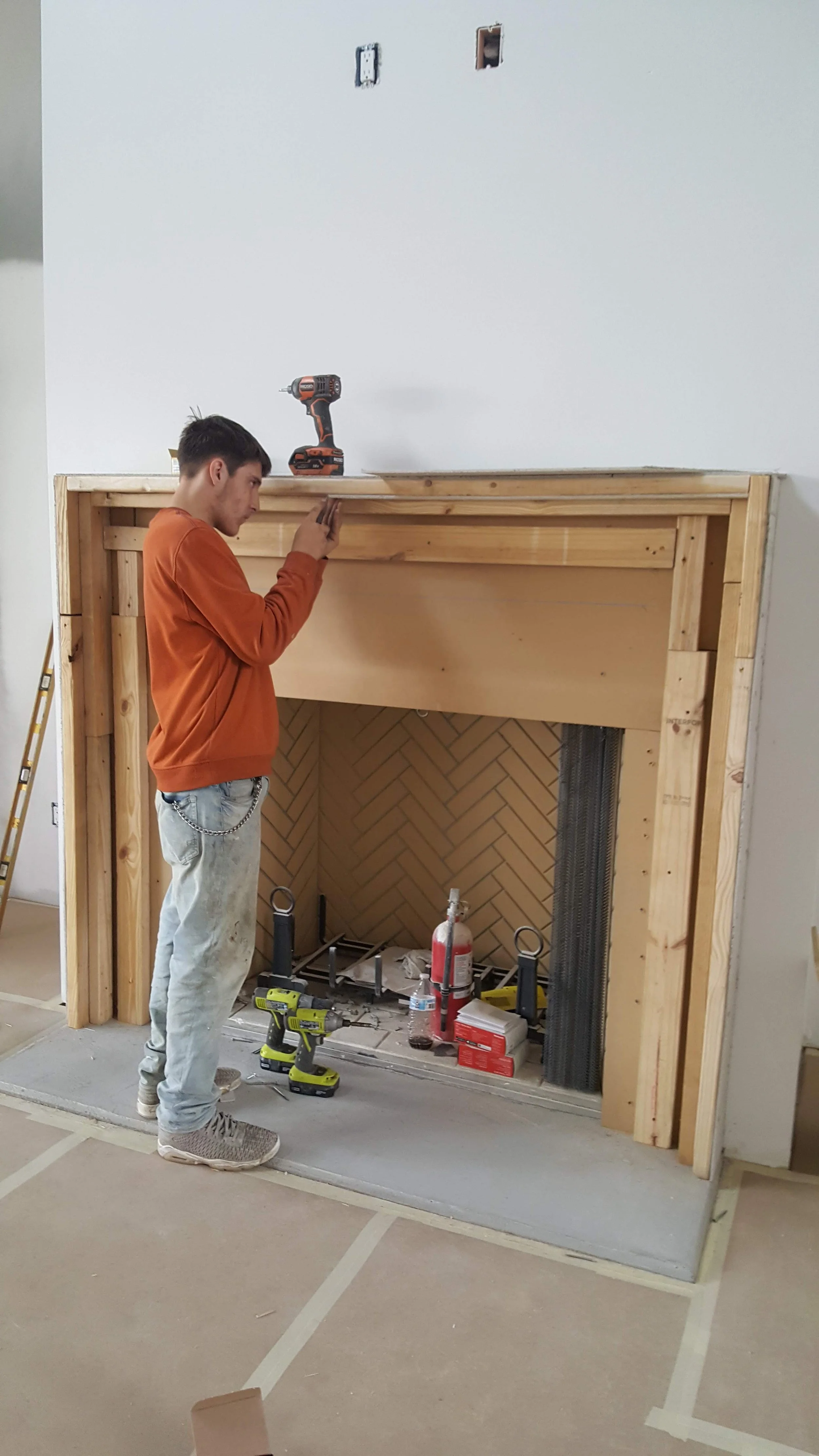 A young man is working on a fireplace construction, standing in front of a partially built wooden frame, with tools and materials around.