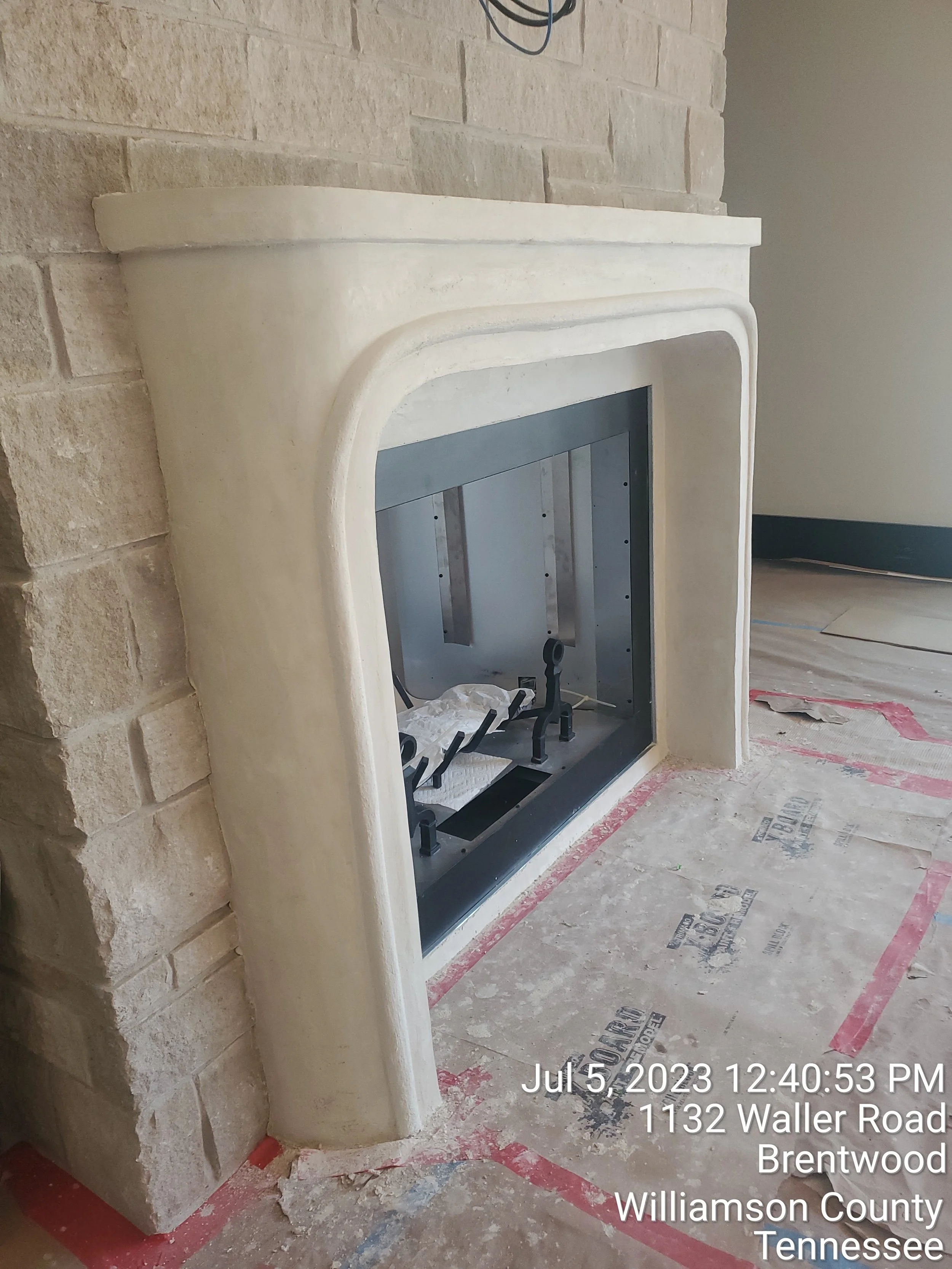 Unfinished fireplace with a beige stucco surround and a black metal frame, located in a room under construction with protective floor covering.