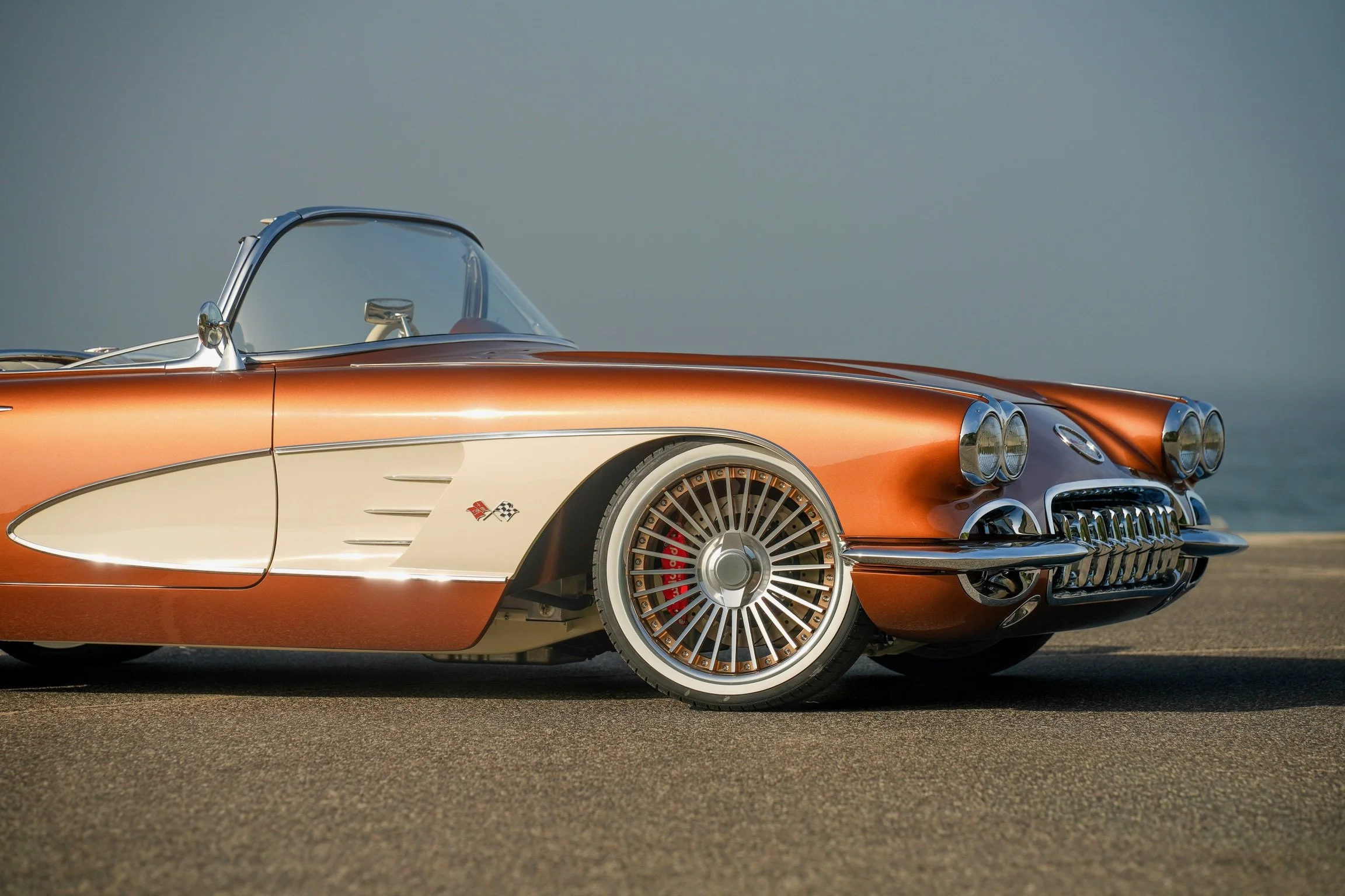 A vintage orange and cream Chevrolet Corvette convertible on a paved surface with a cloudy sky background.