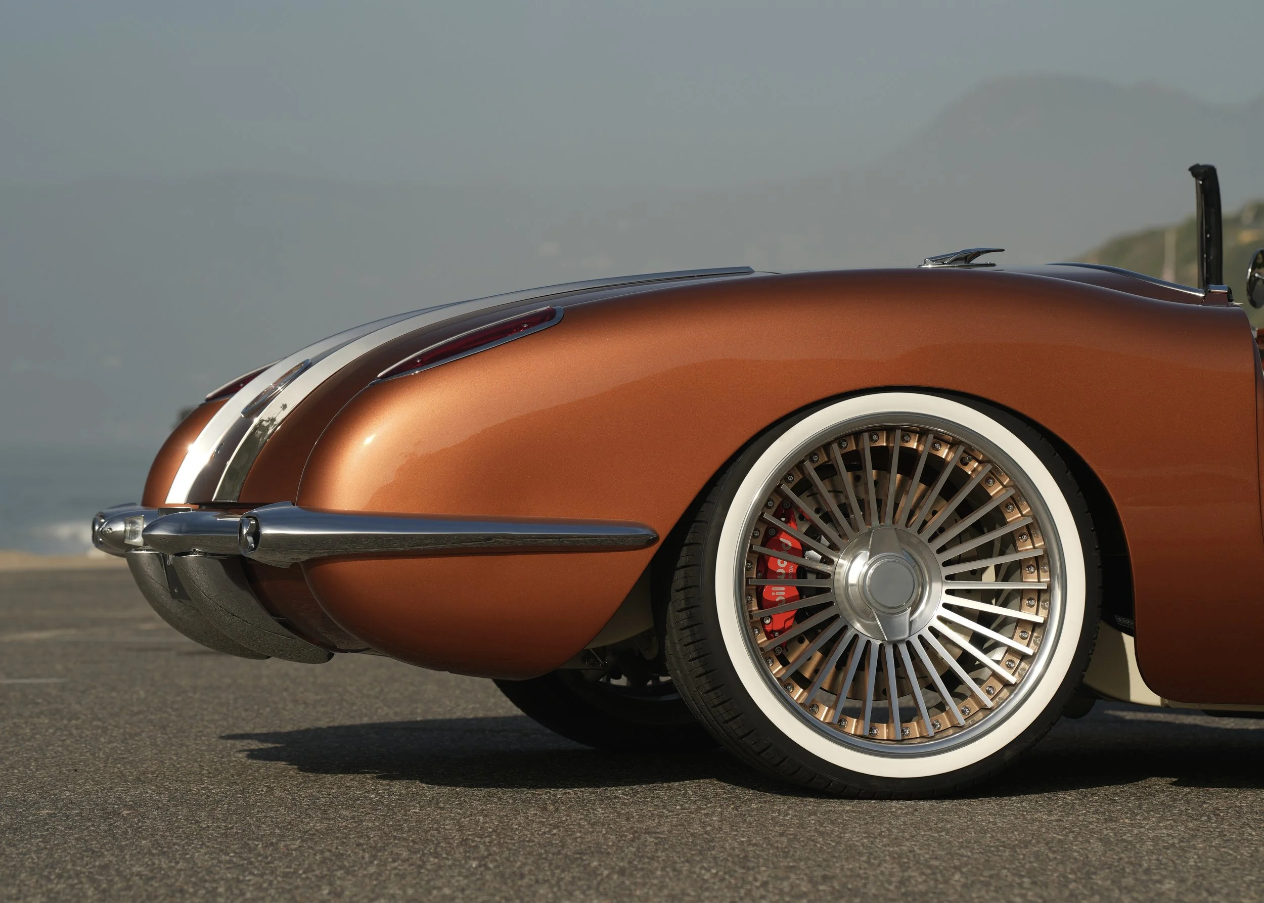 Close-up of the front side of an orange vintage car with wire-spoked wheel and white wall tire, parked on a paved surface with mountains and sky in the background.