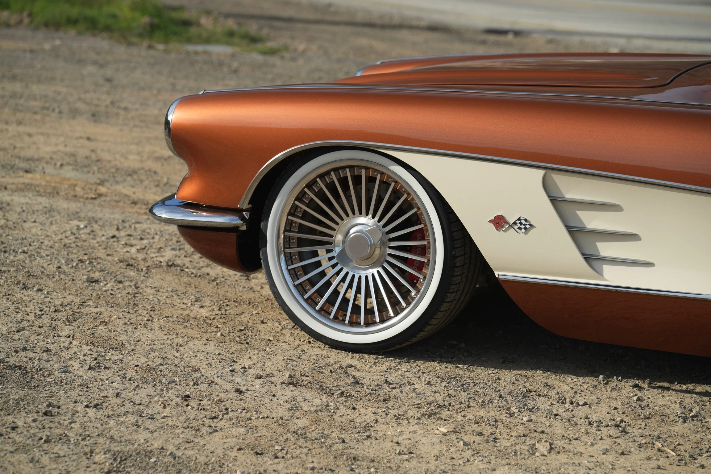 A classic vintage car with a two-tone paint job of metallic copper and cream, featuring a chrome bumper, wire-spoke wheels with whitewall tires, and a small badge with checkered flags on the lower side panel.