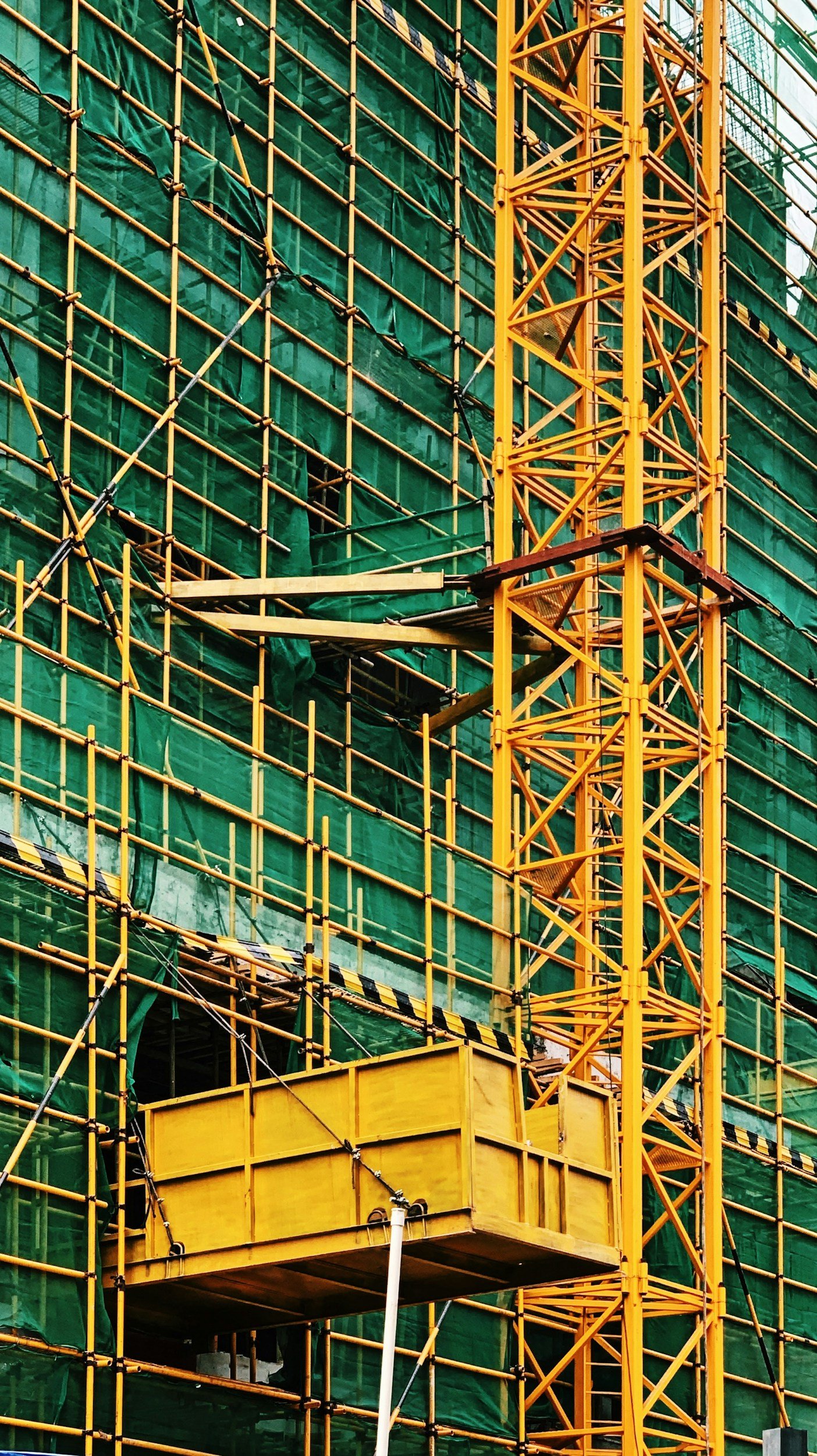 Yellow construction elevator platform on a skyscraper under construction, with green scaffolding and safety netting.